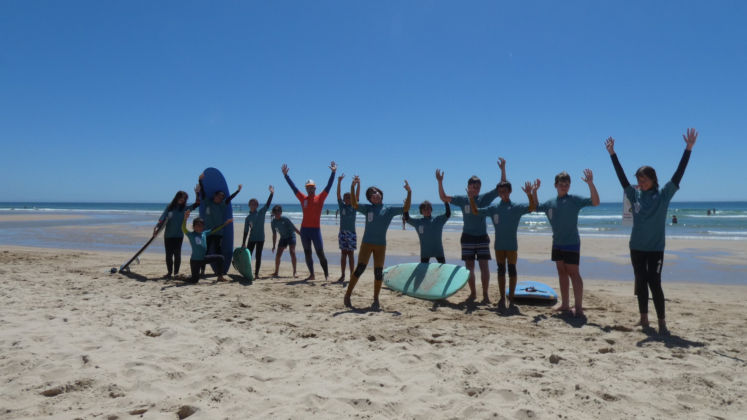 A group of people on a sandy beach at a Time to Surf lesson, standing in a row with surfboards and equipment, raising their arms in the air, with the ocean and clear blue sky in the background.