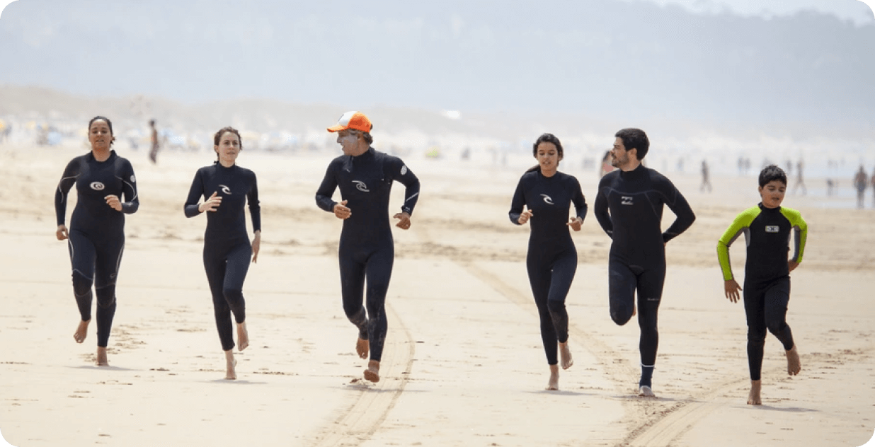 Group of people running on a sandy beach, wearing wetsuits.