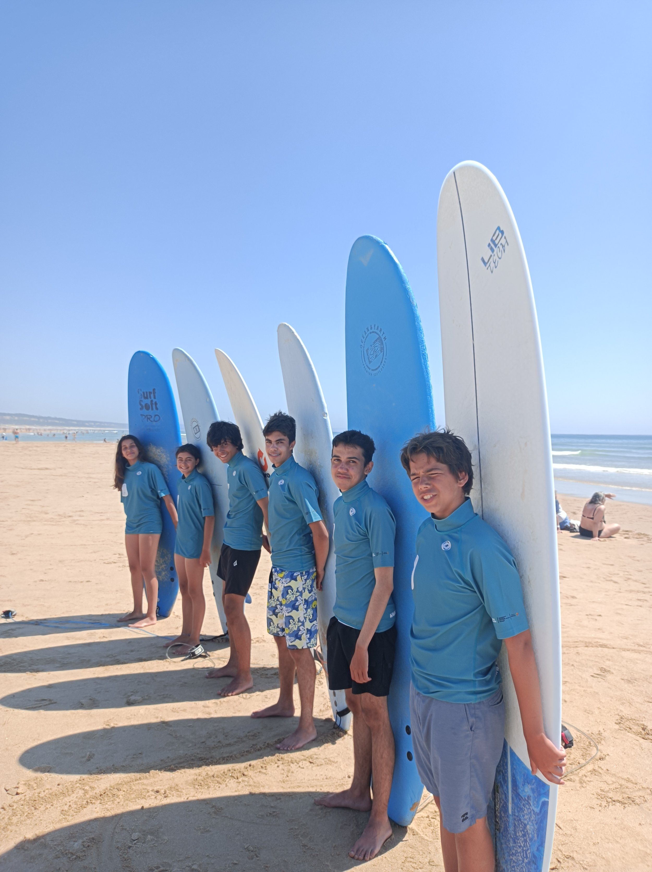 Group of six young people standing on a sandy beach, each holding a different colored surfboard upright behind them, with the ocean and a clear blue sky in the background.