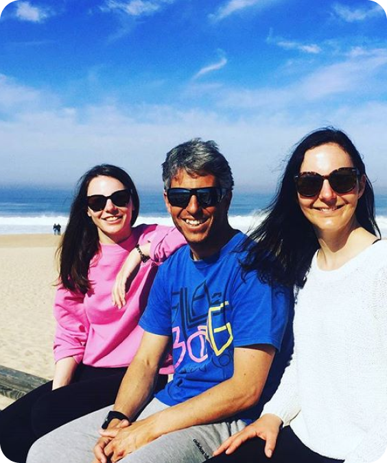 Three friends sitting together on a beach, posing for a photo. They are all wearing sunglasses and smiling. The background shows sand, the ocean, and a blue sky with some clouds.