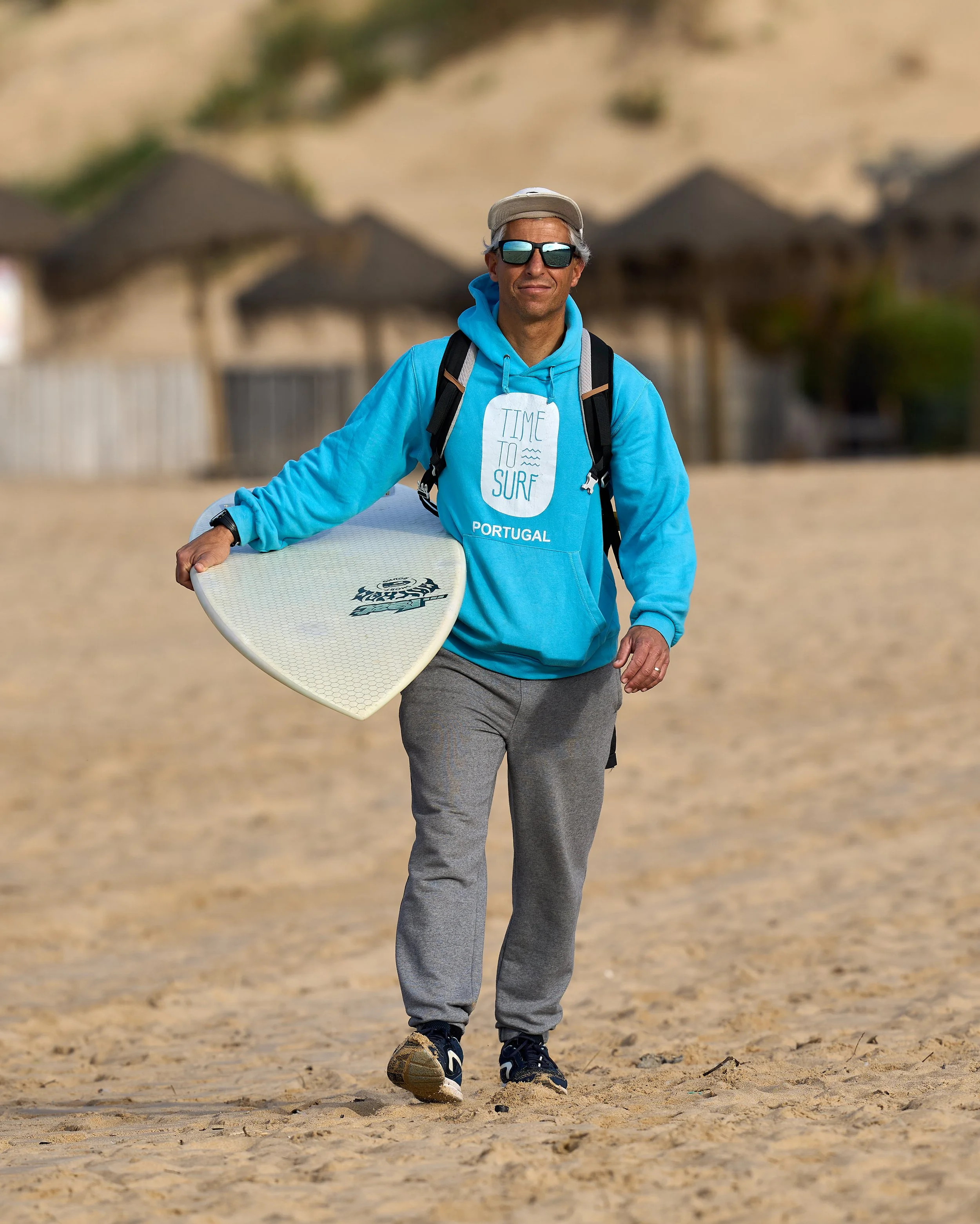 A man walking on a beach holding a surfboard, wearing sunglasses, a beige cap, a bright blue hoodie with 'Time to Surf Portugal' written on it, gray sweatpants, and black sneakers, with beach huts and sandy dunes in the background.