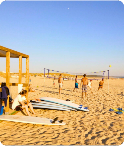 People playing volleyball on a sandy beach with surfboards and a lifeguard tower in the foreground, under a clear blue sky.