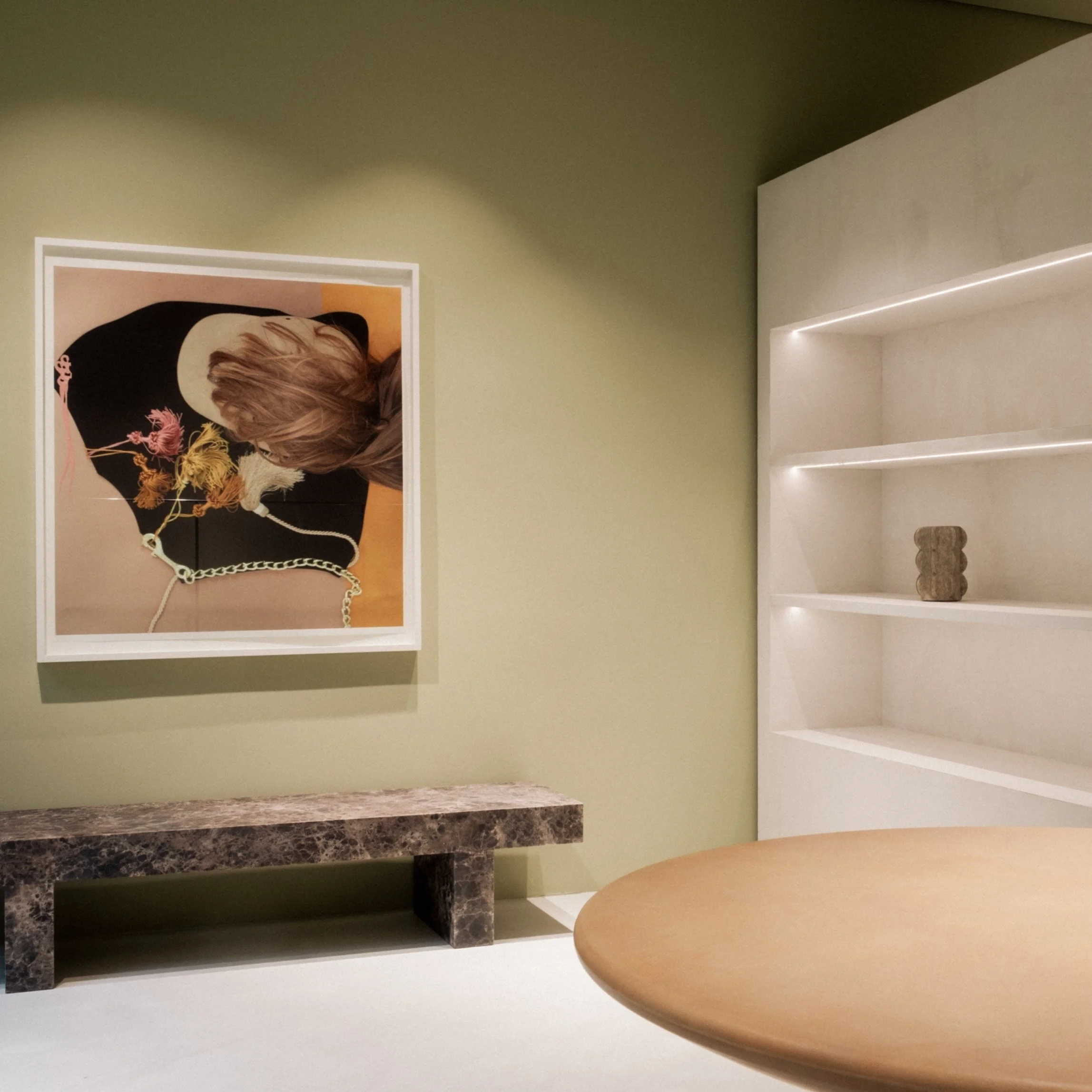 Ein Blick in ein modernes Raum mit grün gestrichener Wand, ein Gemälde eines Frauenporträts, ein brauner Marmortisch und ein weißes Regal mit einer Skulptur.