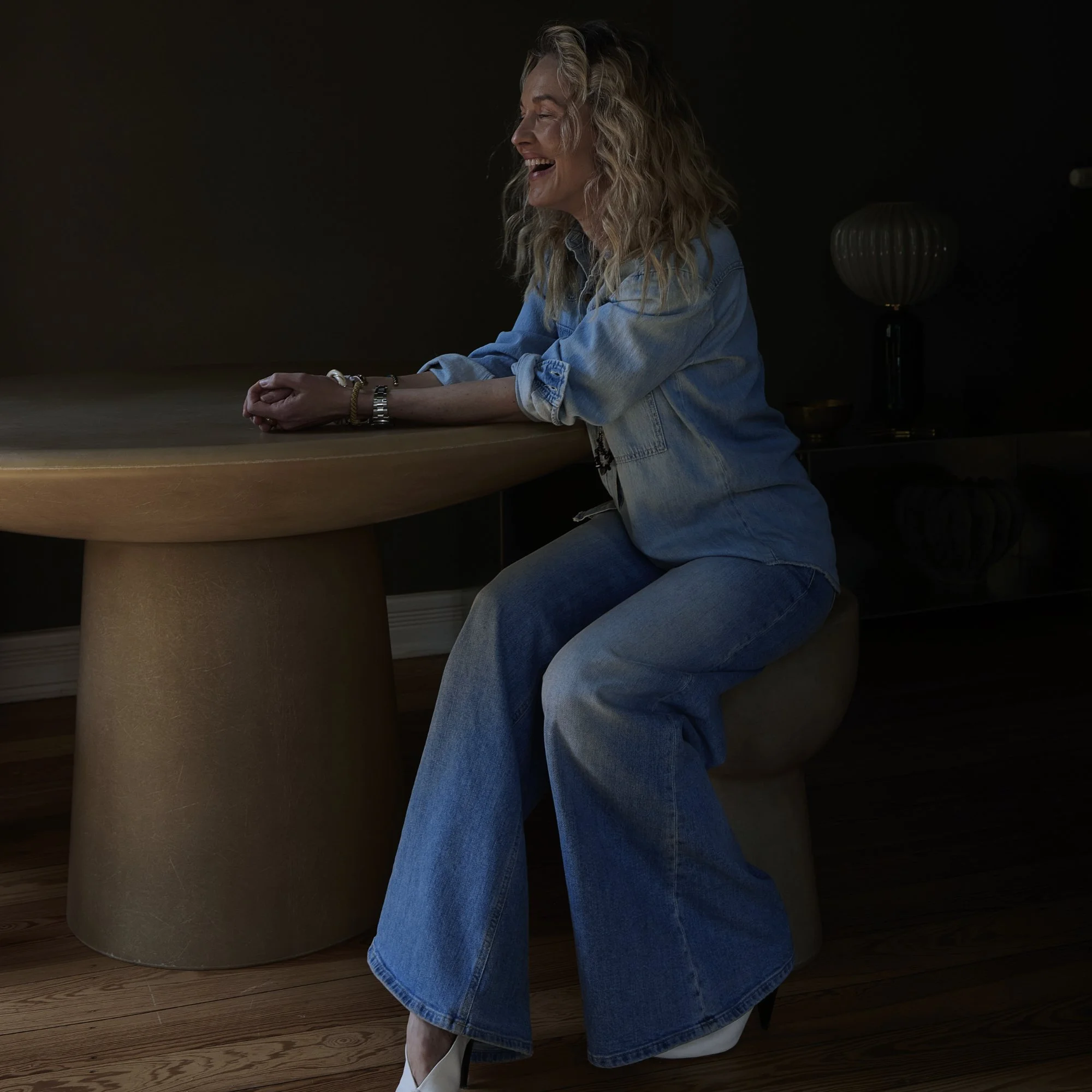 Eine lachende Frau sitzt an einem runden Holztisch in einem dunklen Raum, trägt einen Jeanshemd und Jeans, mit Armreifen und einem Uhrenarmband, ihre Haare sind lockig.