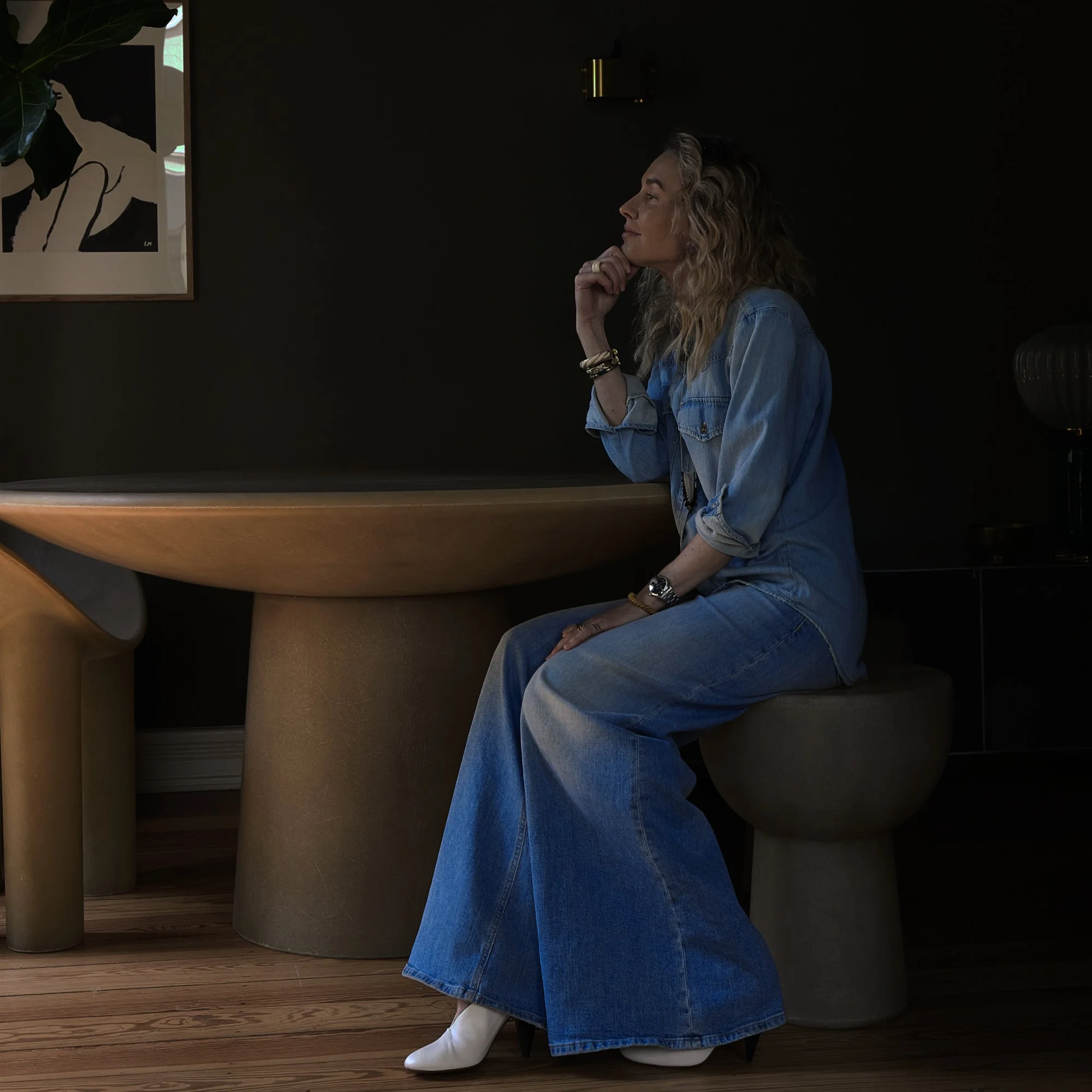 Frau sitzend an einem runden Tisch in einem dunklen Raum, trägt Jeans, White Boots und sieht nachdenklich aus.