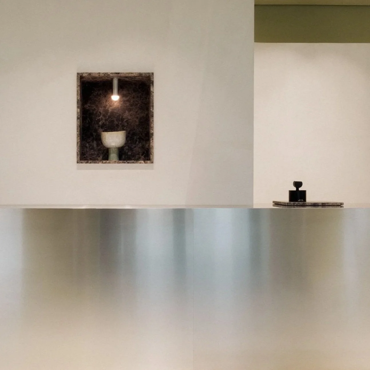 Ein minimalistischer Raum mit einer weißen Wand, einem schwarzen Tisch und einer kunstvollen Nische mit einer weißen Vase und einer kleinen Lampe.