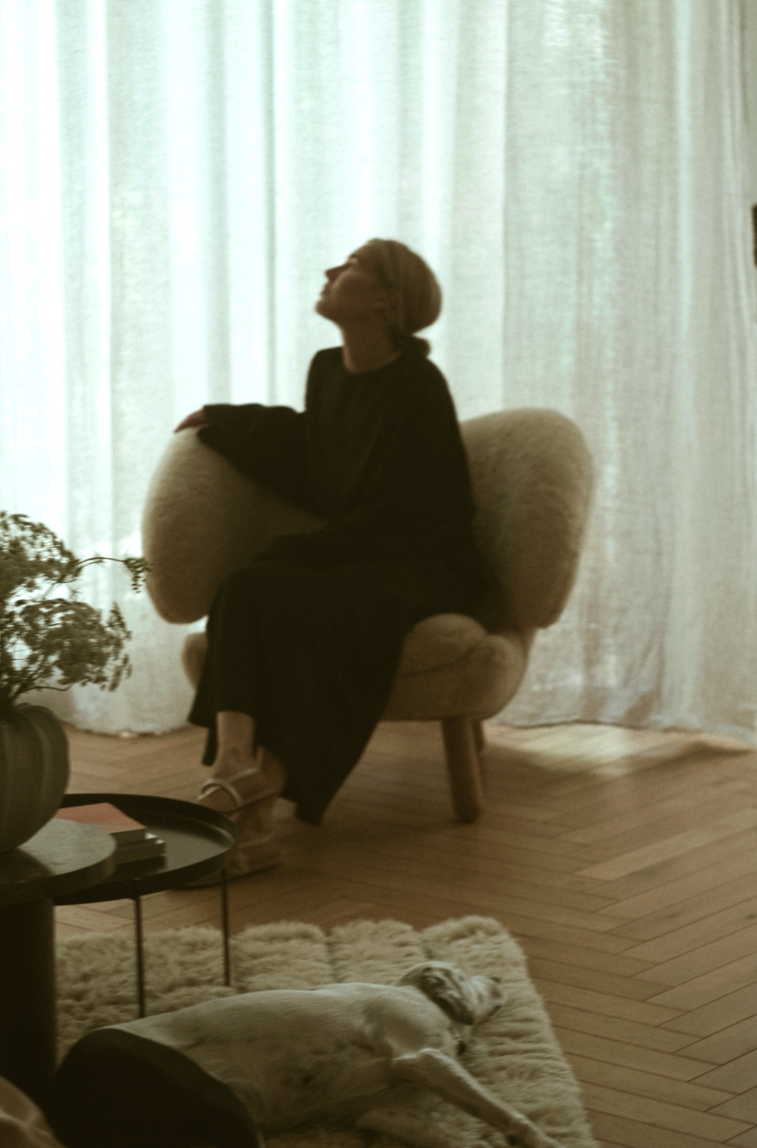Eine Frau sitzt entspannt in einem bequemen Sessel vor einer hellen Gardine, während ein Hund auf einem weichen Teppich davor liegt.