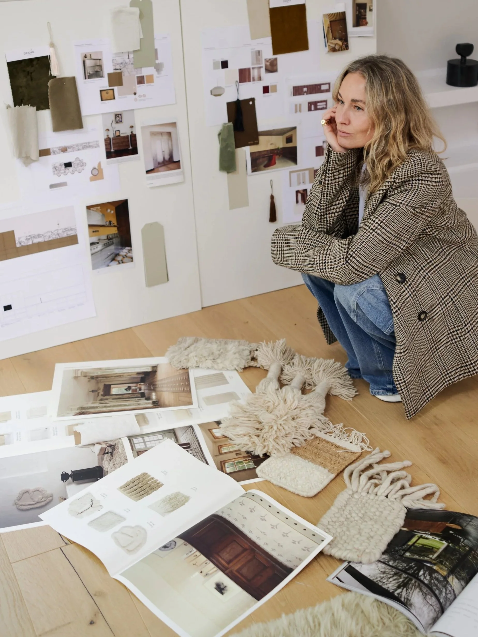 Eine Frau sitzt vor Wand mit Design- und Farbmustern, umgeben von Moodboards, Stoffmustern und Innendesign-Materialien in einem Raum für Inneneinrichtung.