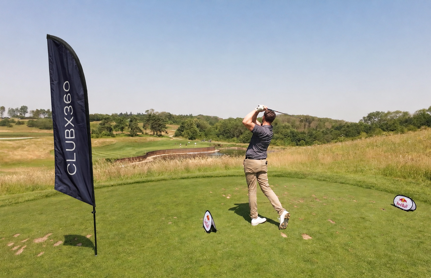 A golfer practicing on a golf course with a club in a swing pose, grass and trees in the background, red bull and club flags on the green.