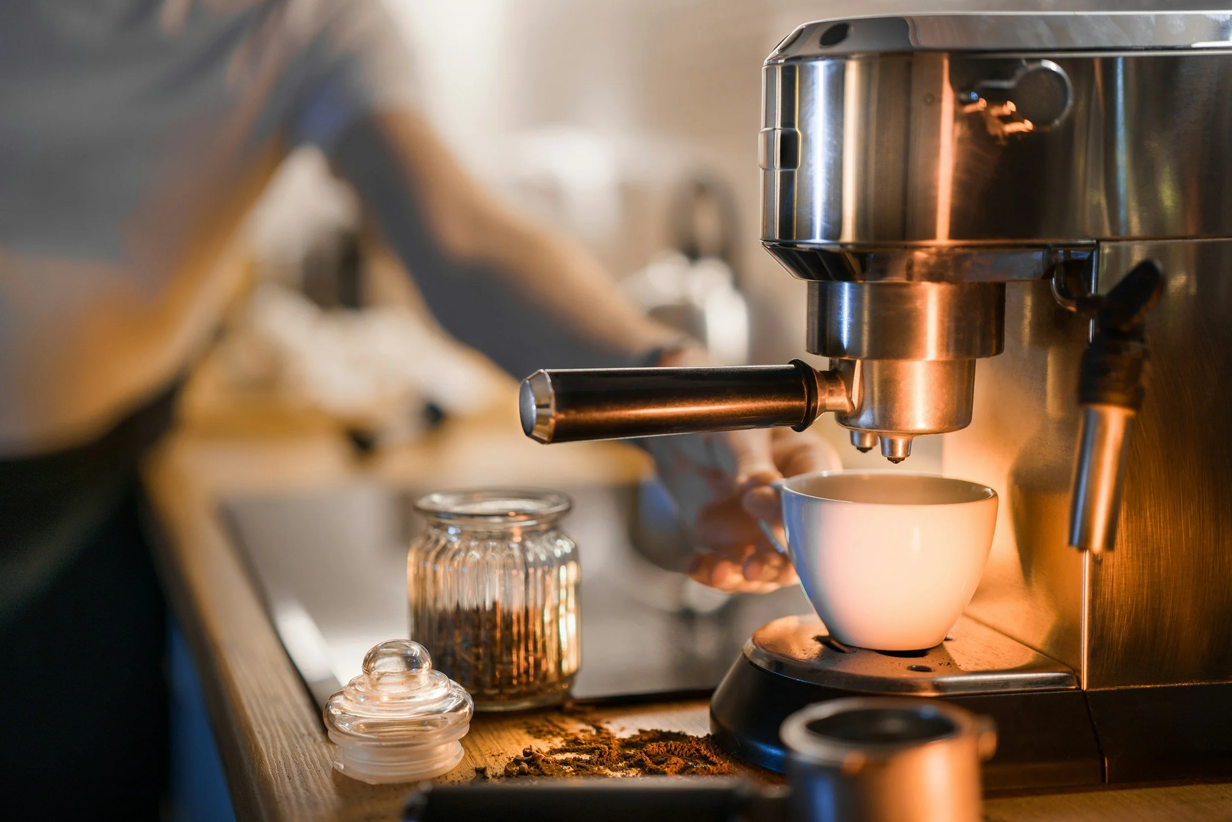 A person preparing coffee with an espresso machine, with a white coffee cup underneath the dispenser, on a wooden counter with coffee beans and a small jar.