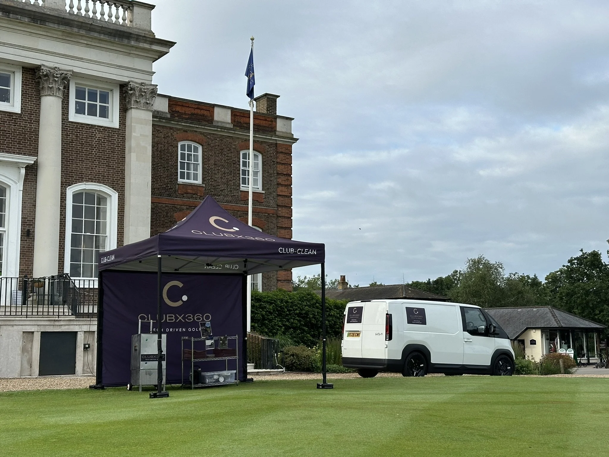 A white van parked on grass in front of a building with a brick facade and white decorative columns. There is a purple canopy with the text 'CLUBX360' and a flagpole with a flag, and another vehicle is partially visible behind the van. The sky is ove
