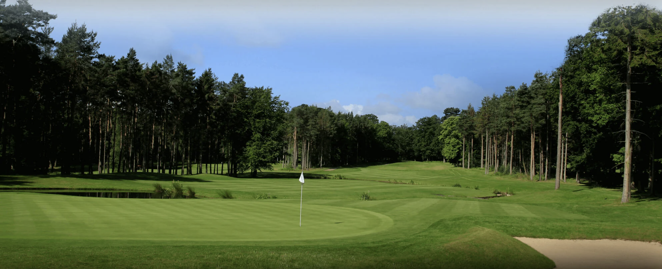 A golf course with a putting green, flagstick, sand trap, and surrounded by trees and a blue sky with some clouds.