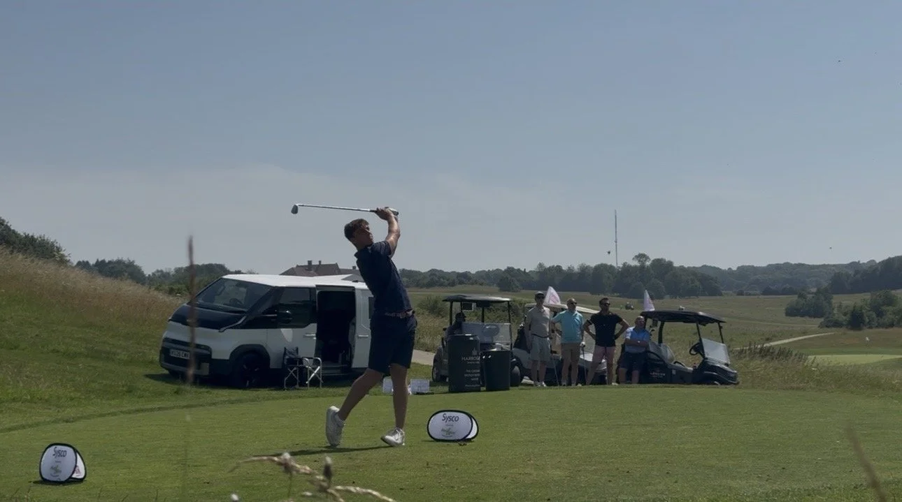 A man swings a golf club on a golf course, with a group of people watching, parked cars, and golf carts in the background under a clear blue sky.
