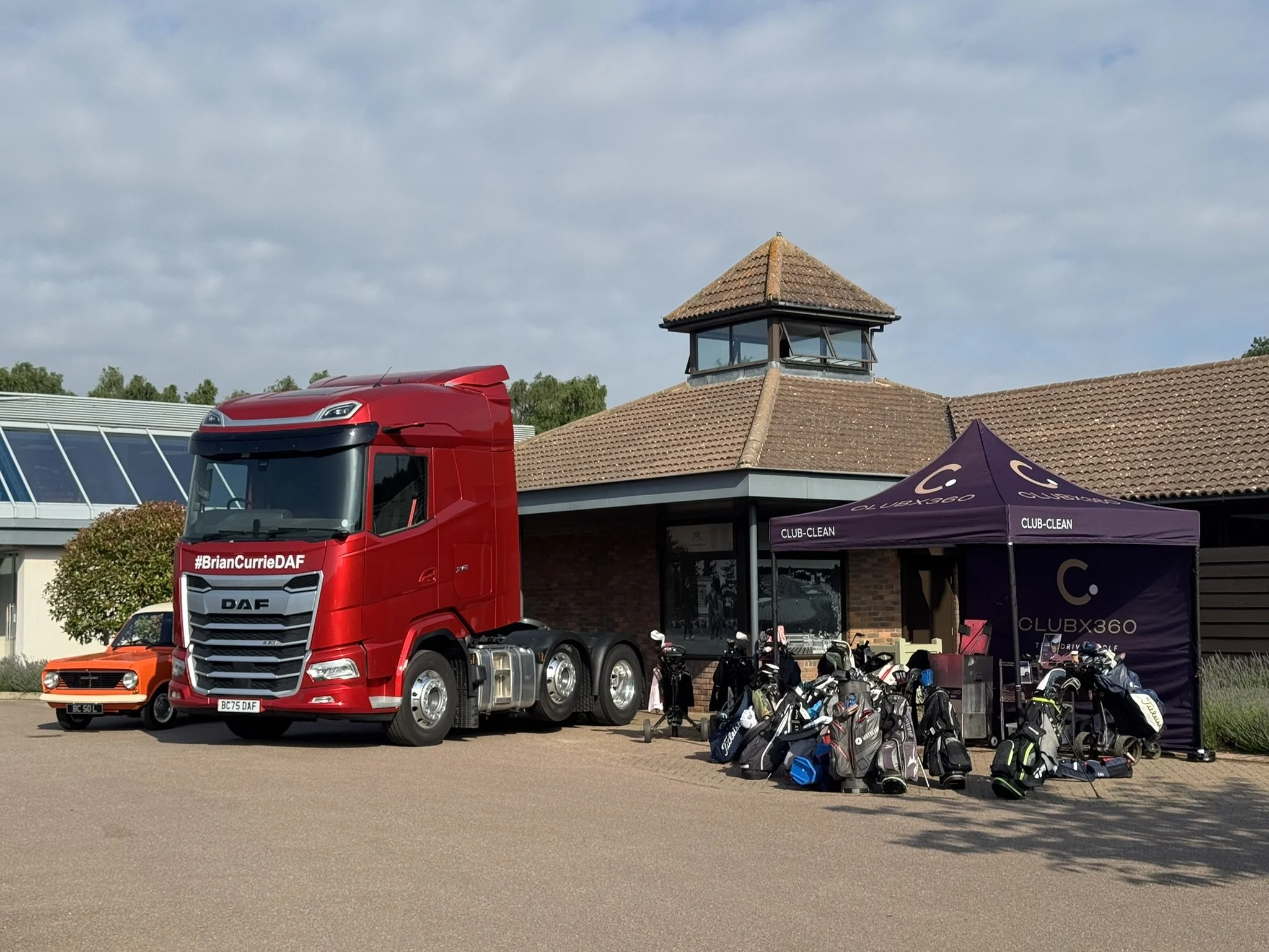 A red semi-truck with the hashtag #BrianCurrieDAF parked outside a building with a stepped roof and a small tower. There are golf bags and carts in the foreground, and a purple canopy with the text 'CLUB-CLEAN' covering some equipment.
