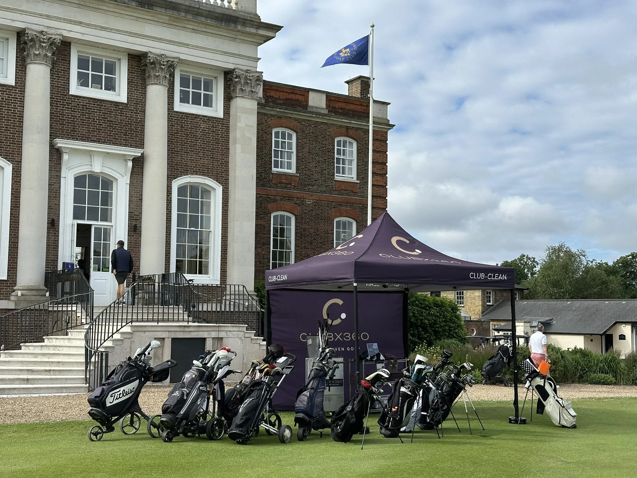 Golf bags and clubs set up on a grassy area near a building with a flag flying overhead and a purple tent labeled 'Club Clean'. People are in the background near the building's entrance and on the grounds.