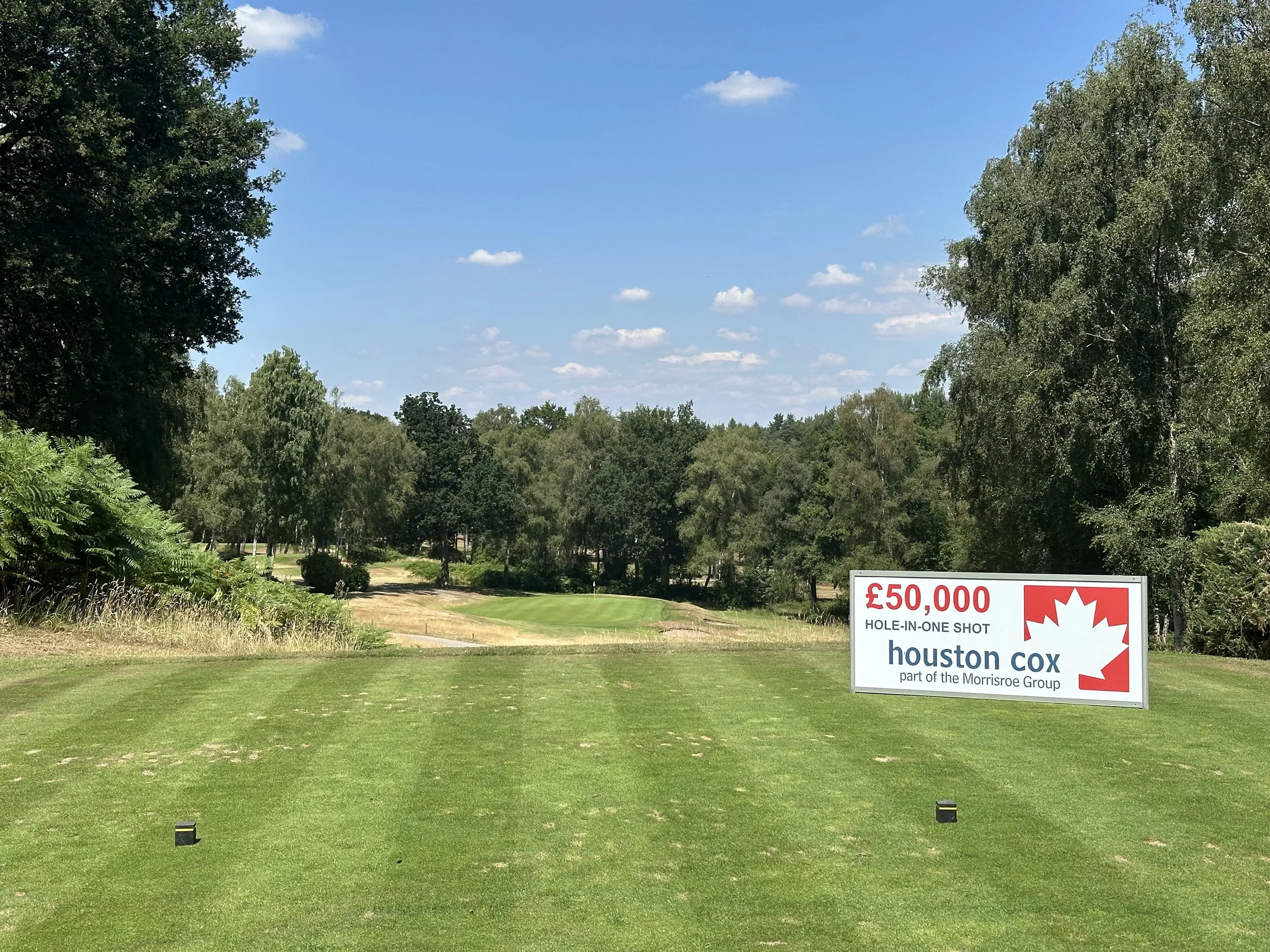 A golf course with a green, trees on the sides, and a sign that reads "£50,000 HOLE-IN-ONE SHOT Houston Cox part of the Morrisroe Group".