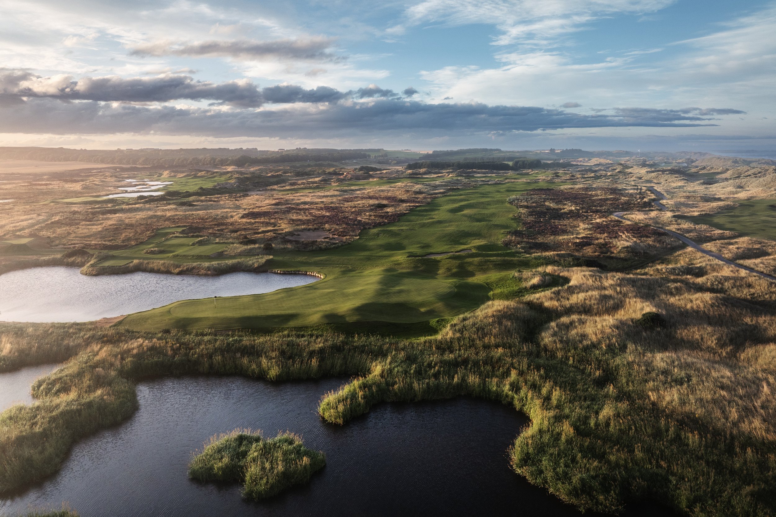 Aerial view of a golf course surrounded by ponds and grassy hills, under a partly cloudy sky.