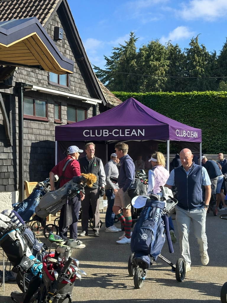 People next to a purple tent with 'CLUB-CLEAN' written on it, some with golf bags and clubs, at a golf course or club, clear blue sky in the background.