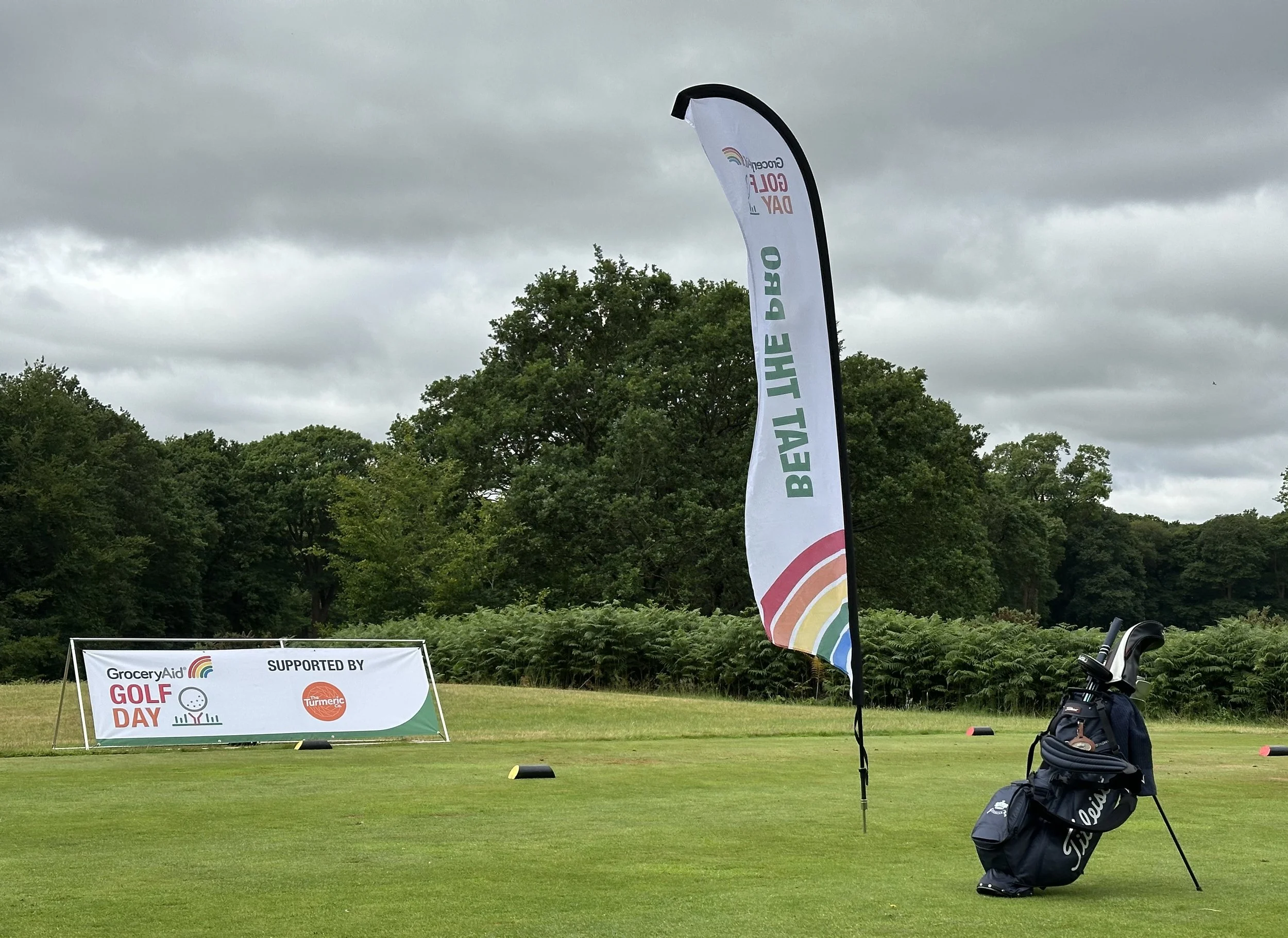 A golf course with promotional flags and signs for an event called 'GOLF DAY' supported by GroceryAid and Turmeric. There is a stand of golf clubs and a golf bag on the grass, amidst cloudy skies.