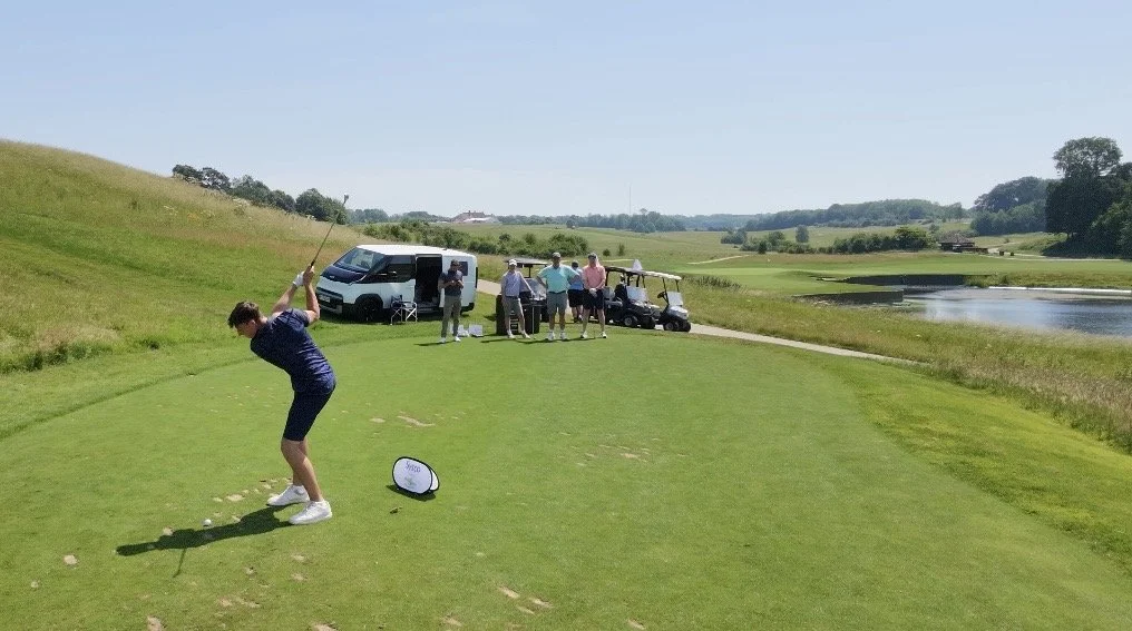 Person in a blue shirt and shorts swinging a golf club on a golf course next to water, with several people and vehicles in the background.