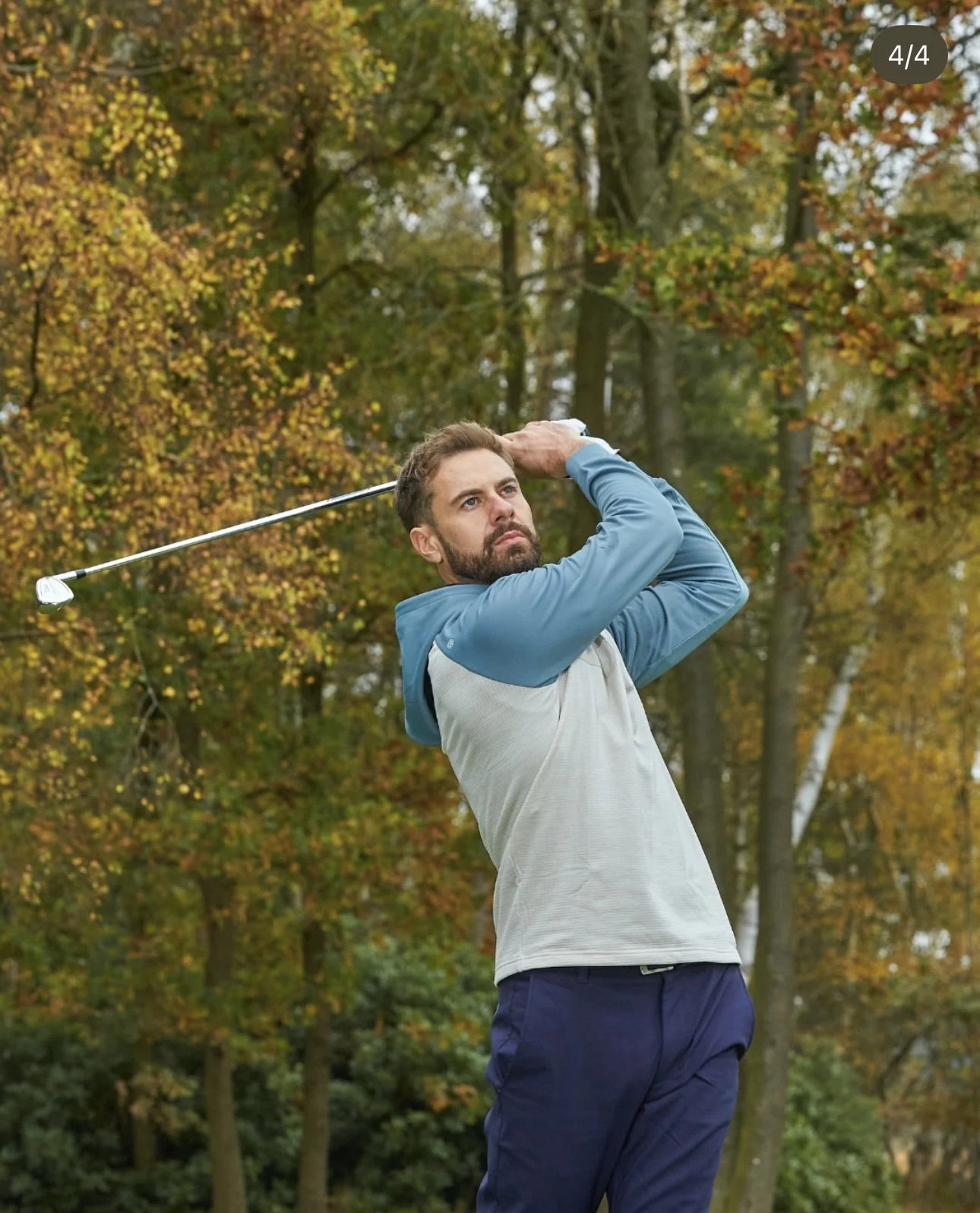 A man playing golf, mid-swing, outdoors with trees in autumn colors in the background.