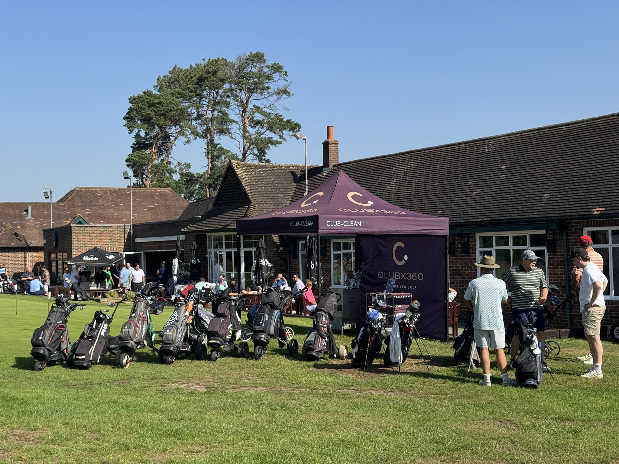 A group of people gather at a golf event on a sunny day, with golf bags and equipment set up on a grassy area in front of a building with a tent and trees in the background.