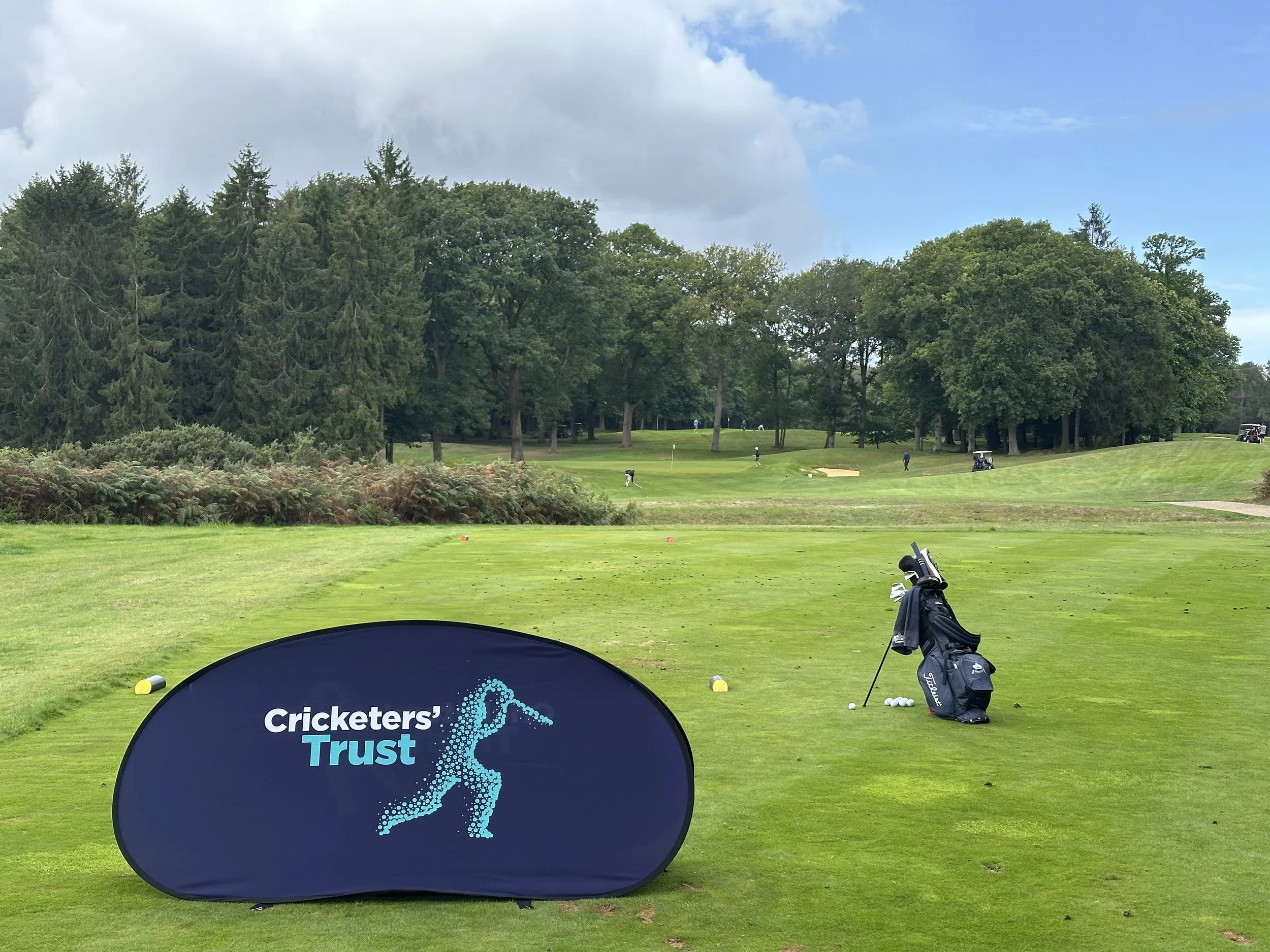 Golf course with trees in the background, golf carts, and people playing golf. A Cricketers Trust sign is in the foreground.