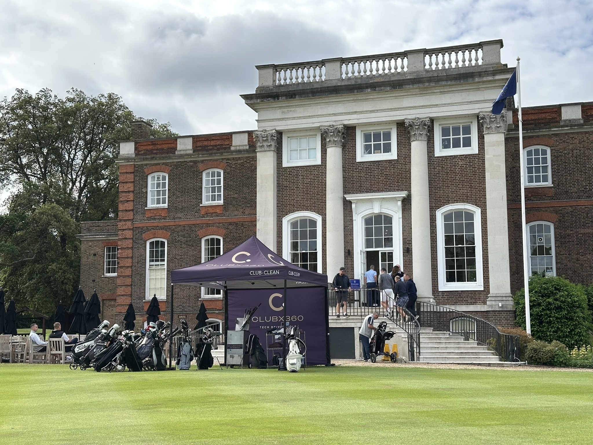 A large brick building with white decorative columns and a balcony on top, located on a golf course. People are near the entrance, and there are golf carts parked nearby. A purple tent with the logo 'CLUBX360' is set up in front.