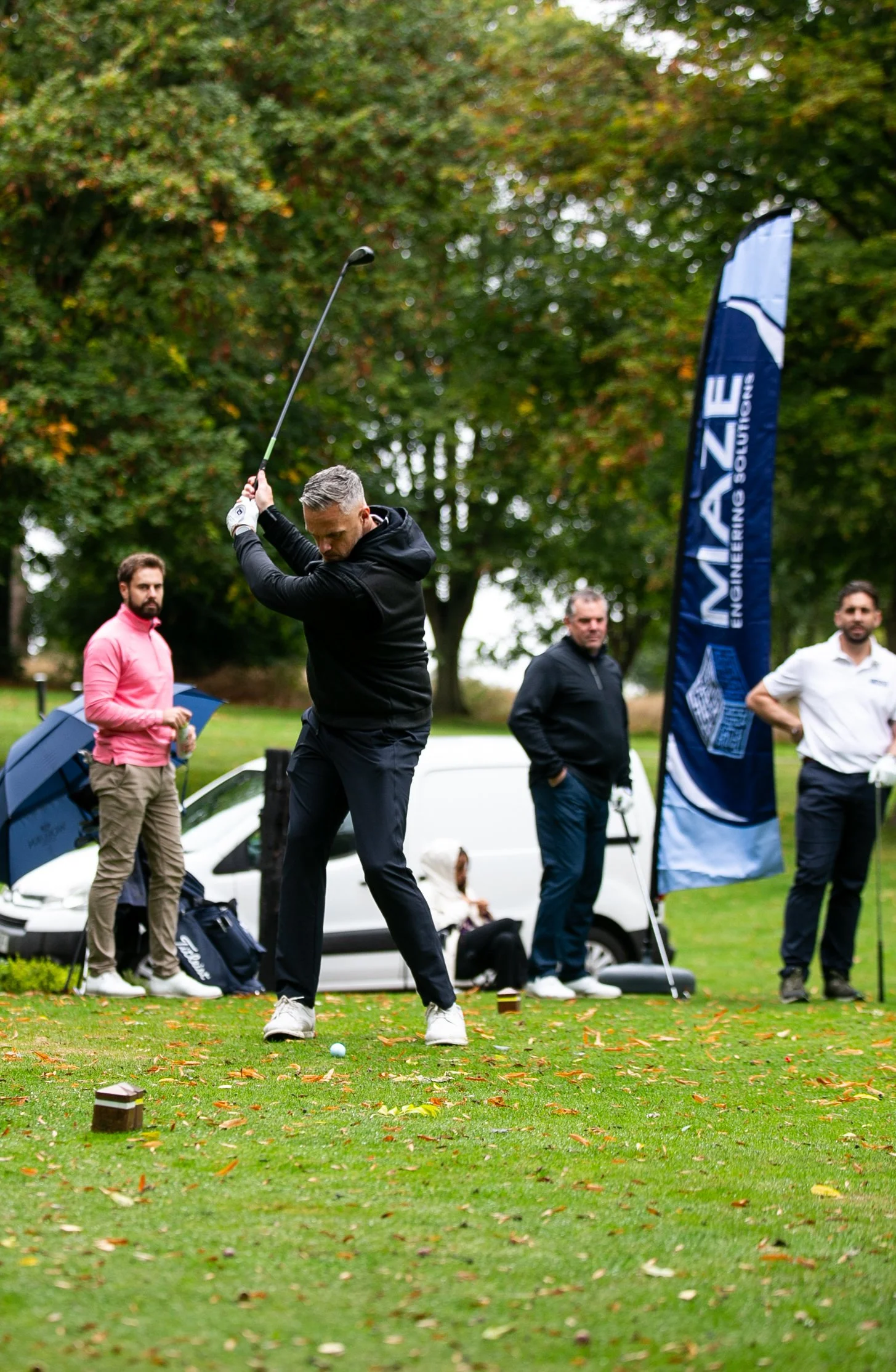 A man in black golf attire swinging a golf club, with three other men watching on a golf course with fall leaves and a banner that reads "MAIZE ENGINEERING SOLUTIONS."