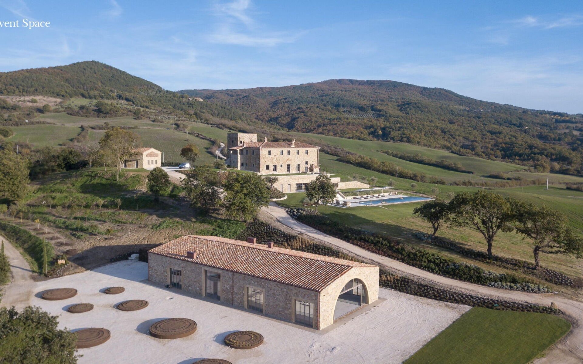 Aerial view of a rural estate with stone buildings, a swimming pool, and surrounding green fields and hills.