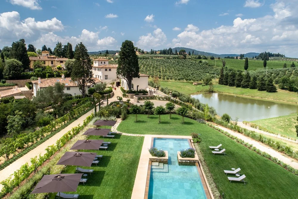 A scenic view of a luxurious mansion with a pool, surrounded by greenery, vineyards, and a lake, with rolling hills in the background under a partly cloudy sky.