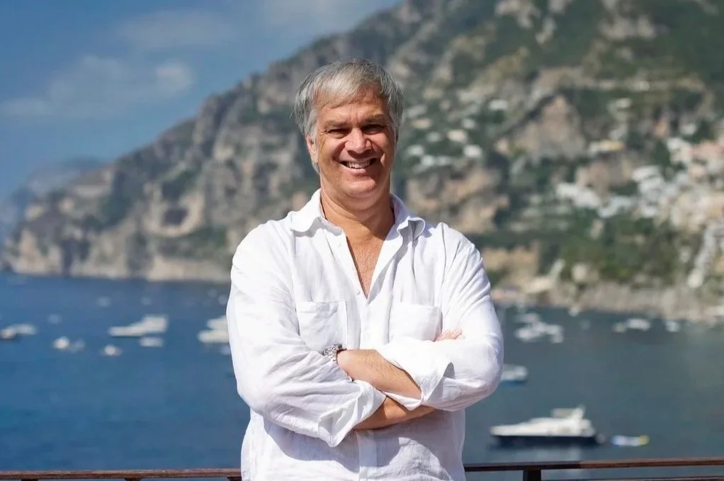 A smiling man with gray hair and a white shirt standing with arms crossed outdoors, with a view of a large body of water, boats, and steep cliffs or mountains in the background.