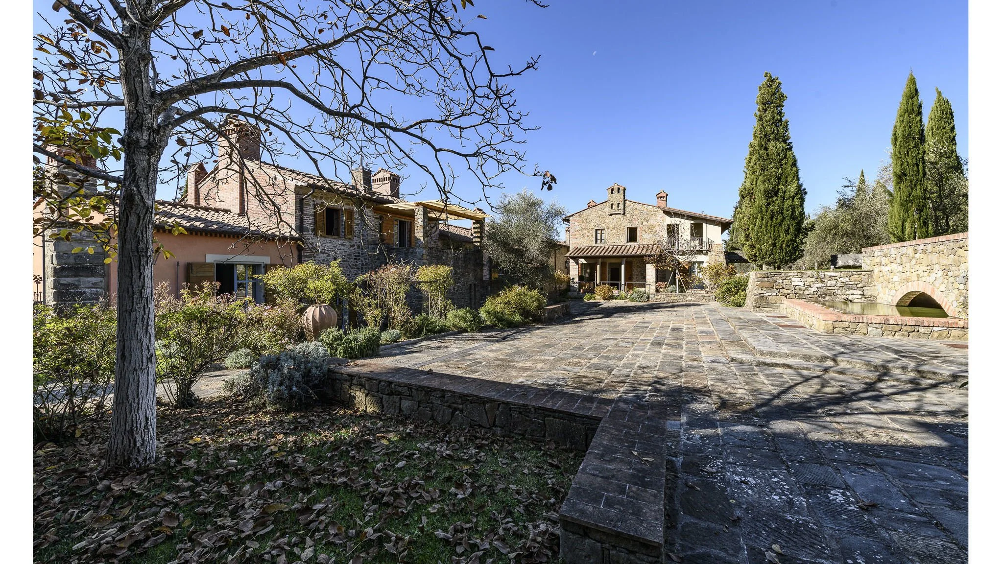 An outdoor view of a rustic house with stone and stucco walls, surrounded by trees and shrubs, on a sunny day with a clear blue sky.