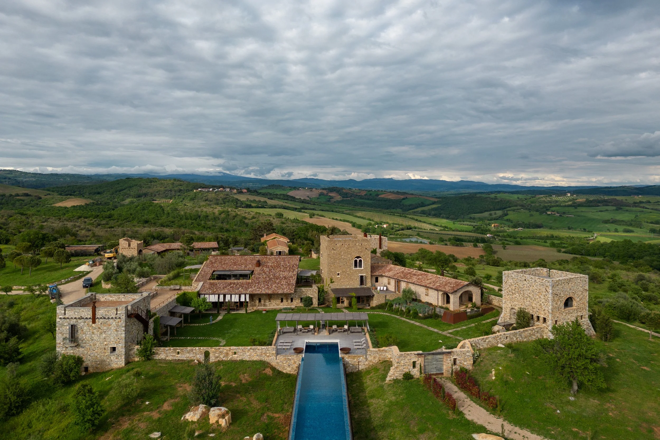 Aerial view of a rustic estate with castle-like stone towers, a swimming pool, and surrounding greenery in a rural landscape under cloudy sky.