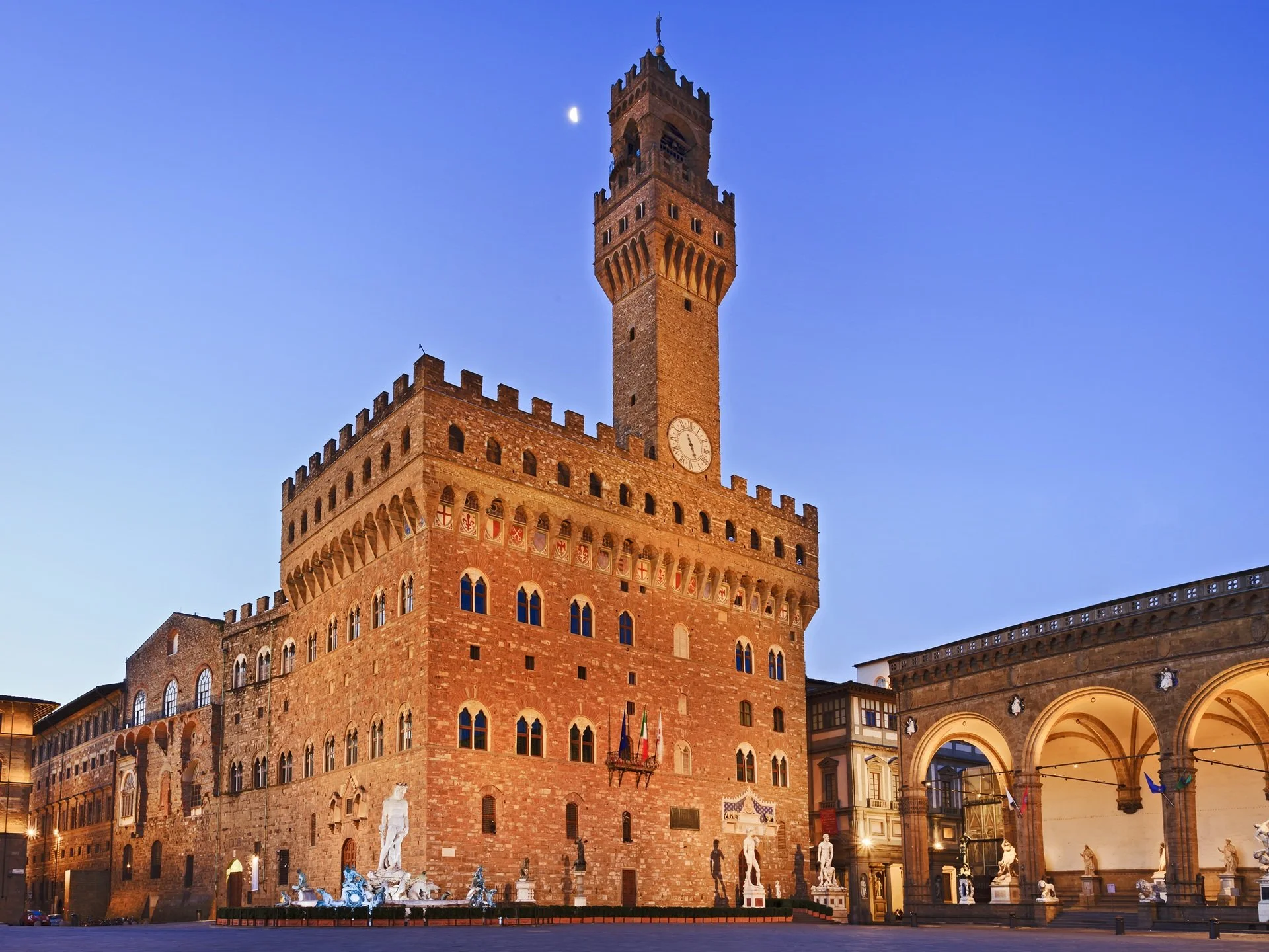 The historic Palazzo Vecchio in Florence, Italy, with statues in front, illuminated at dusk, and the moon visible in the sky.