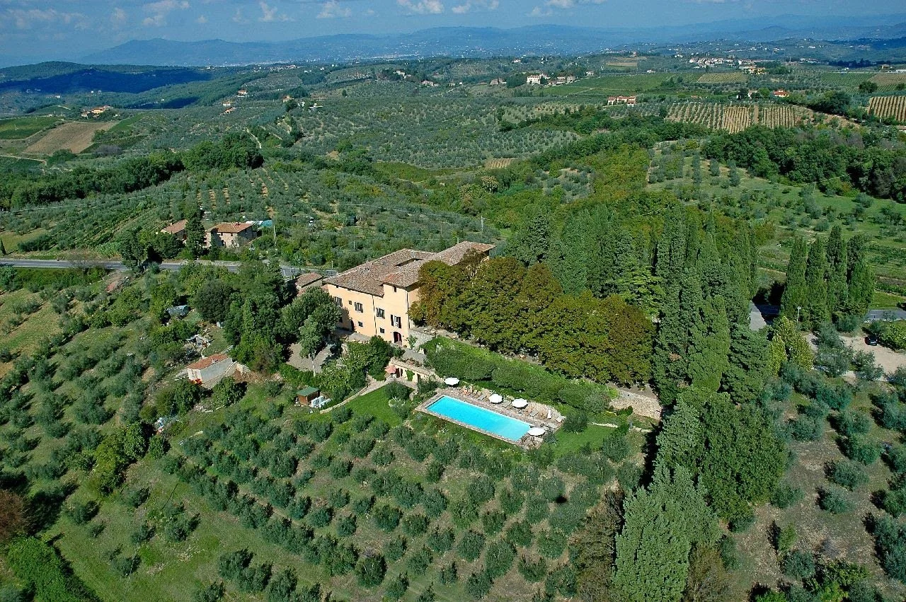 Aerial view of a countryside with a large house, swimming pool, and landscaped garden surrounded by rolling green hills, vineyards, and farmland under a blue sky.