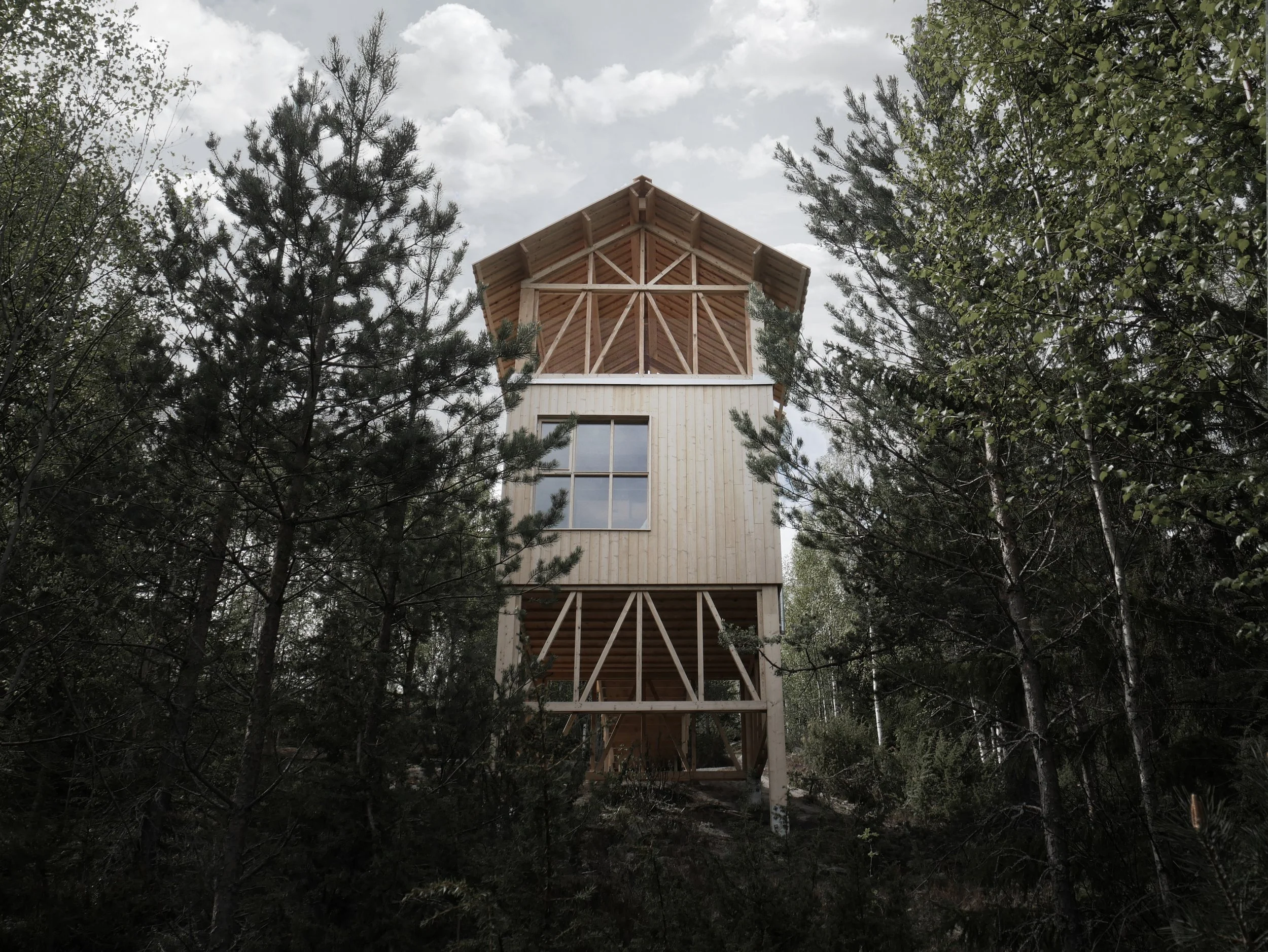 A wooden treehouse built among tall trees with a gable roof and large window, situated in a forested area with a cloudy sky in the background.