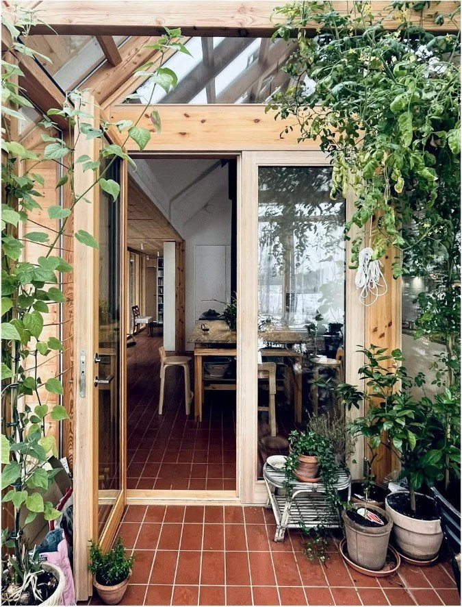 View of an indoor garden or sunroom with potted plants, wooden framing, and glass doors leading into a cozy dining area with a table and chairs inside a home.