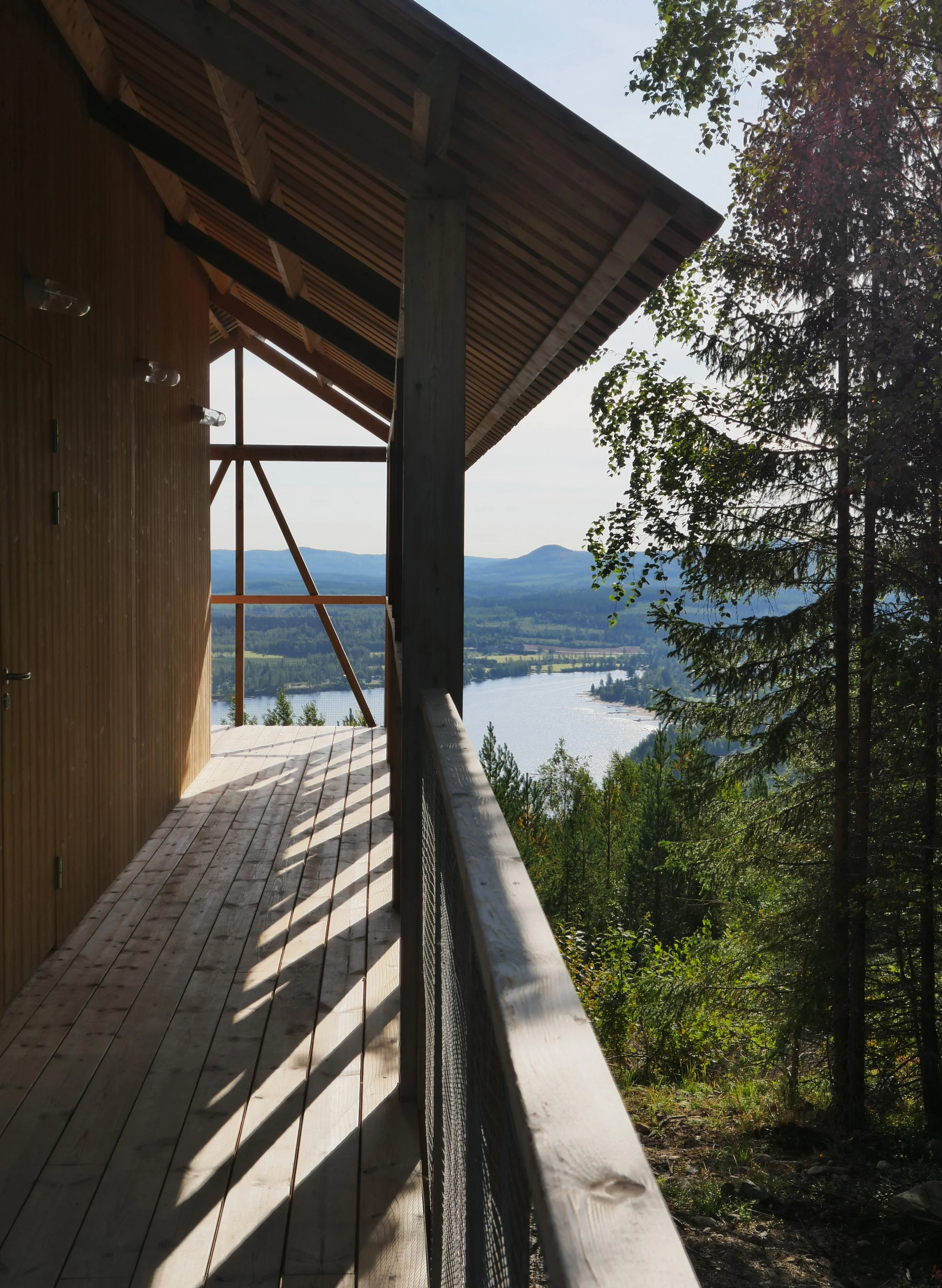 A wooden balcony overlooking a scenic river and distant mountains, surrounded by green trees and clear sky.