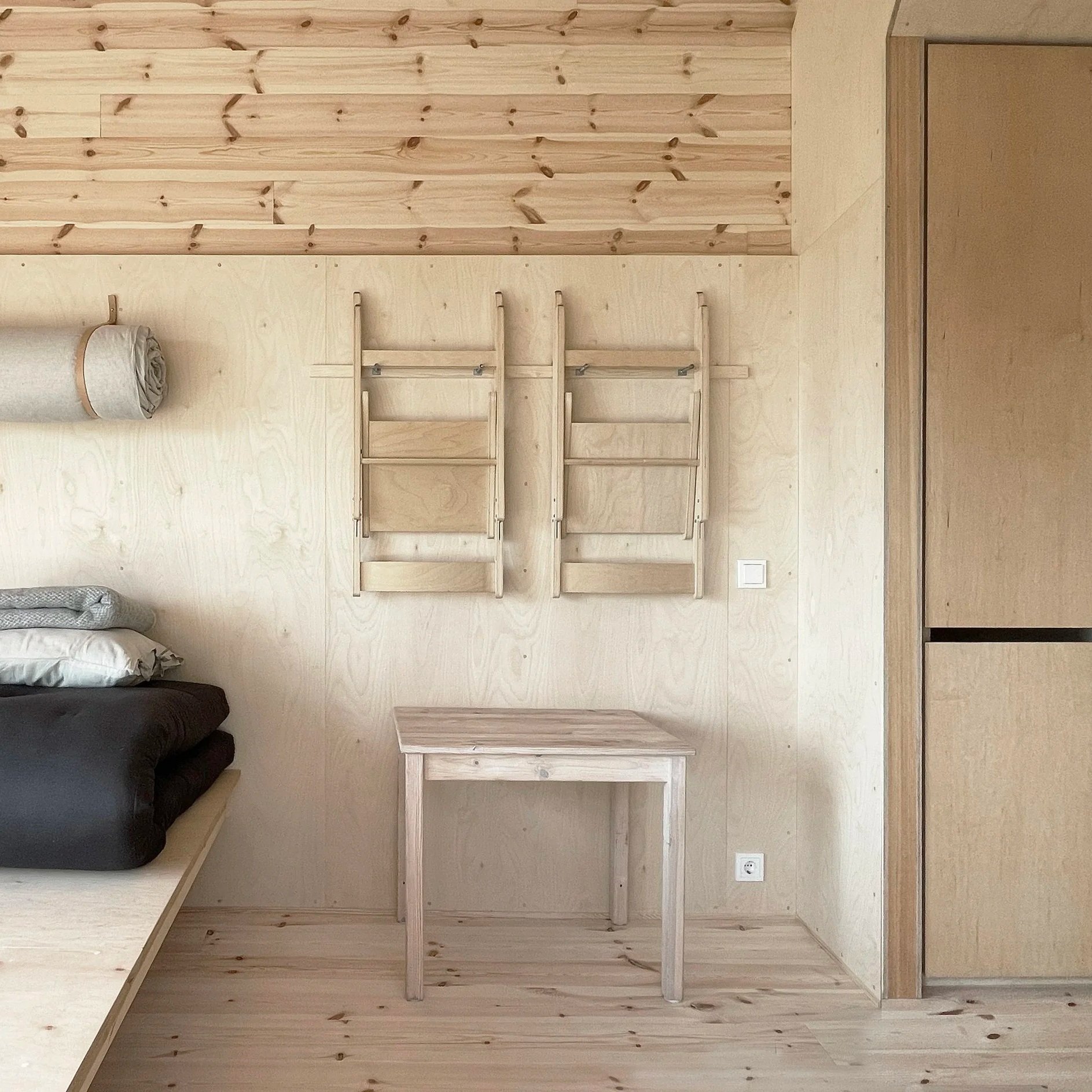 A minimalist wooden room with a bed, a small table, and wall-mounted wooden ladders or racks.
