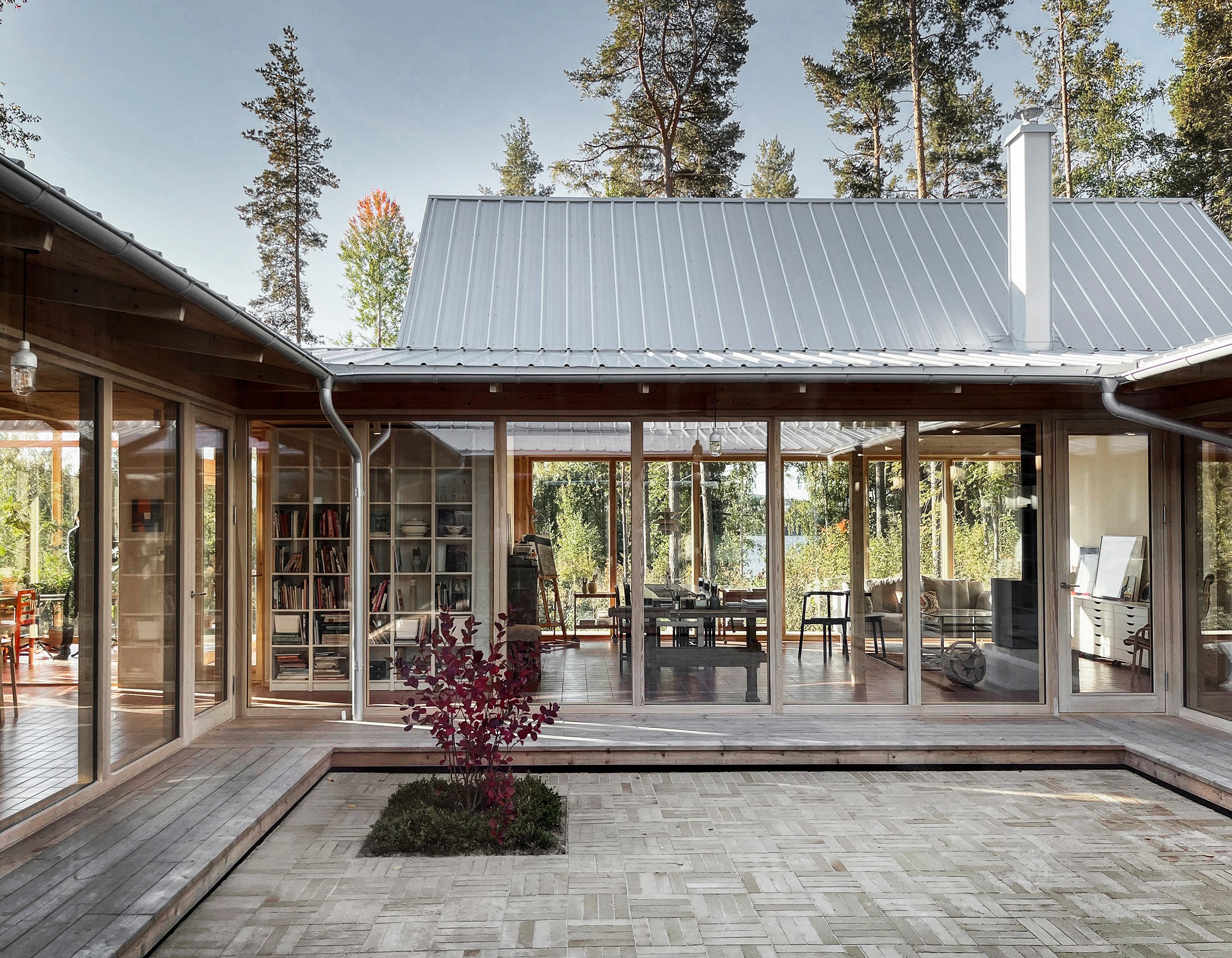 View of a modern house with large glass windows and a metal roof, surrounded by trees, with a small paved courtyard and a flower bed in the foreground.