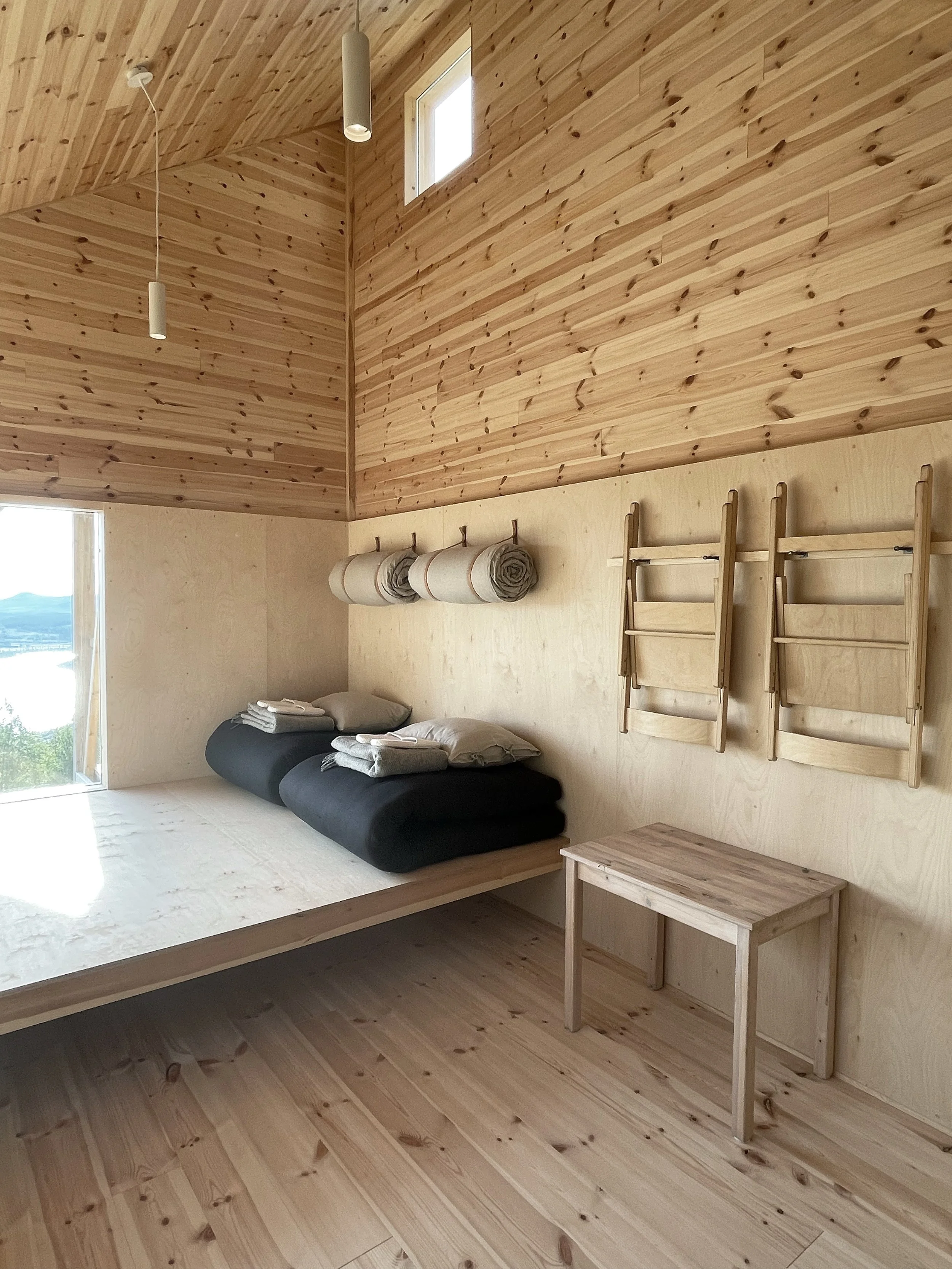 A minimalist bedroom with wooden walls, ceiling, and floor. There is a bed with black bedding, neatly folded towels and pillows on top. Above the bed, three rolled-up blankets are hung on hooks. To the right, two foldable wooden ladders are mounted on the wall, and a small wooden table is placed nearby. A window and small square window provide natural light.