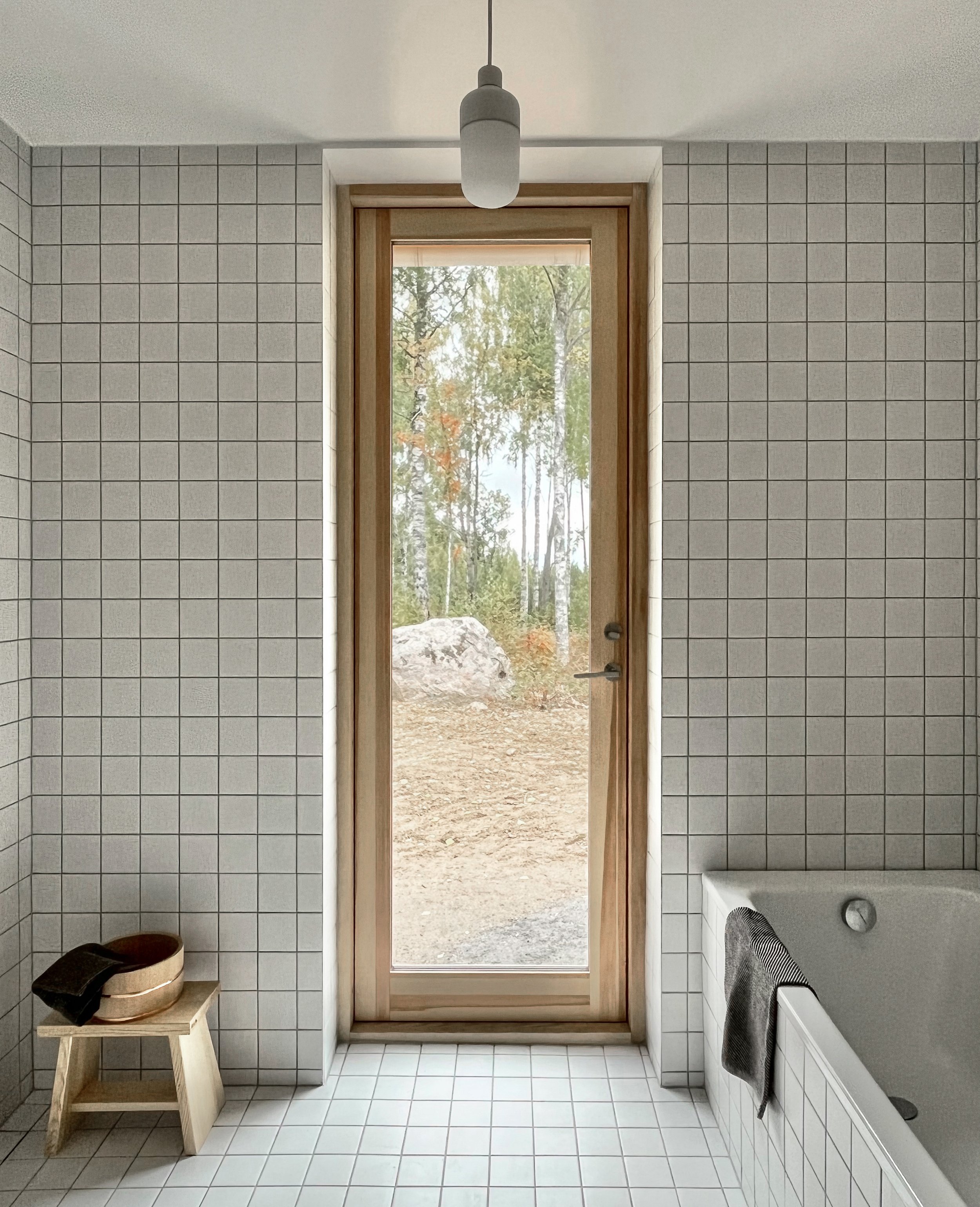 Modern bathroom with white tiled walls and floor, a bathtub with a dark gray towel, a small wooden stool with a bowl and towel, and a large glass door leading outside to a wooded area.