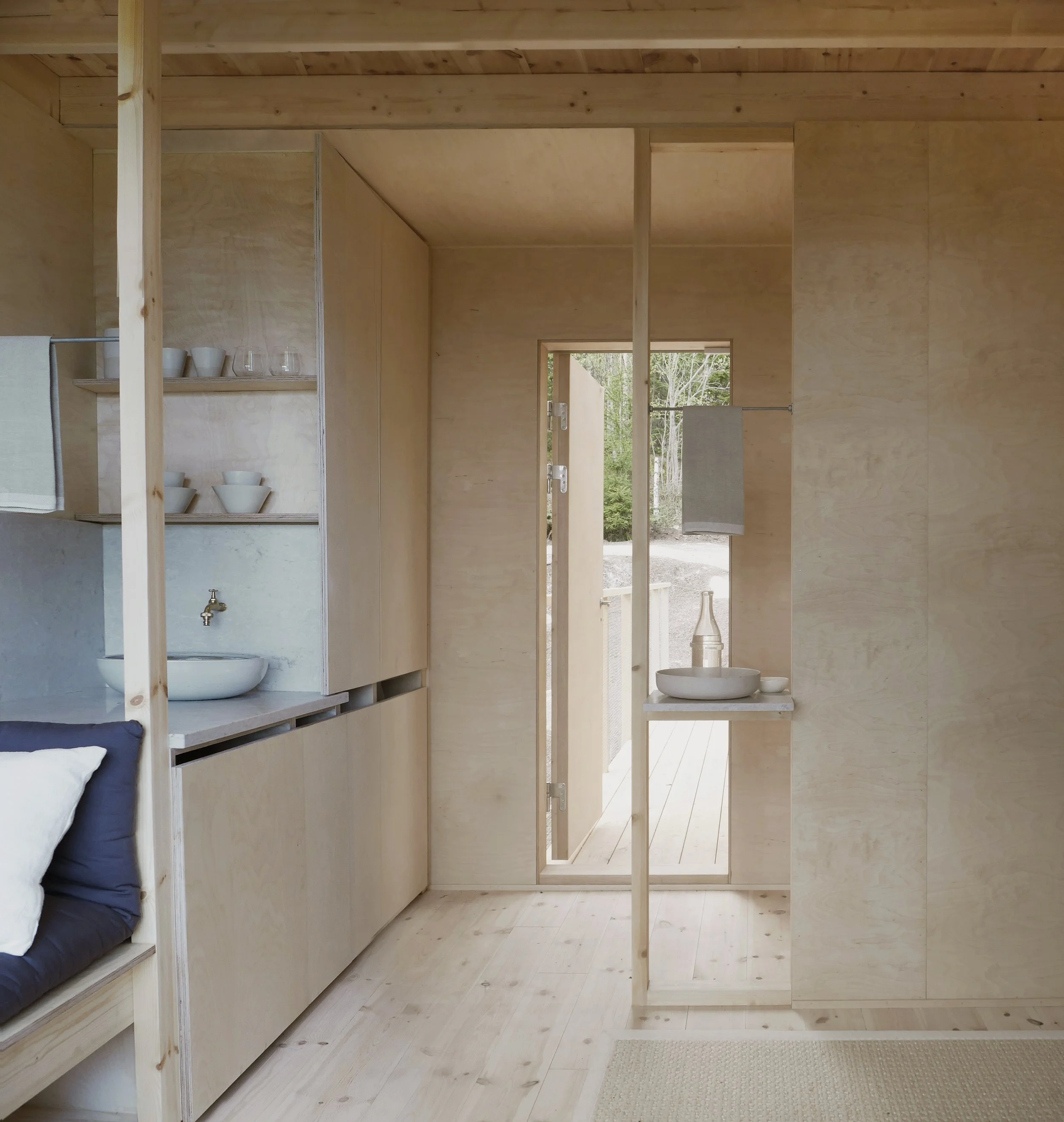 Interior view showing light wood walls, shelves with white bowls and glasses, a small sink with a faucet, and a door opening to an outdoor area with trees.