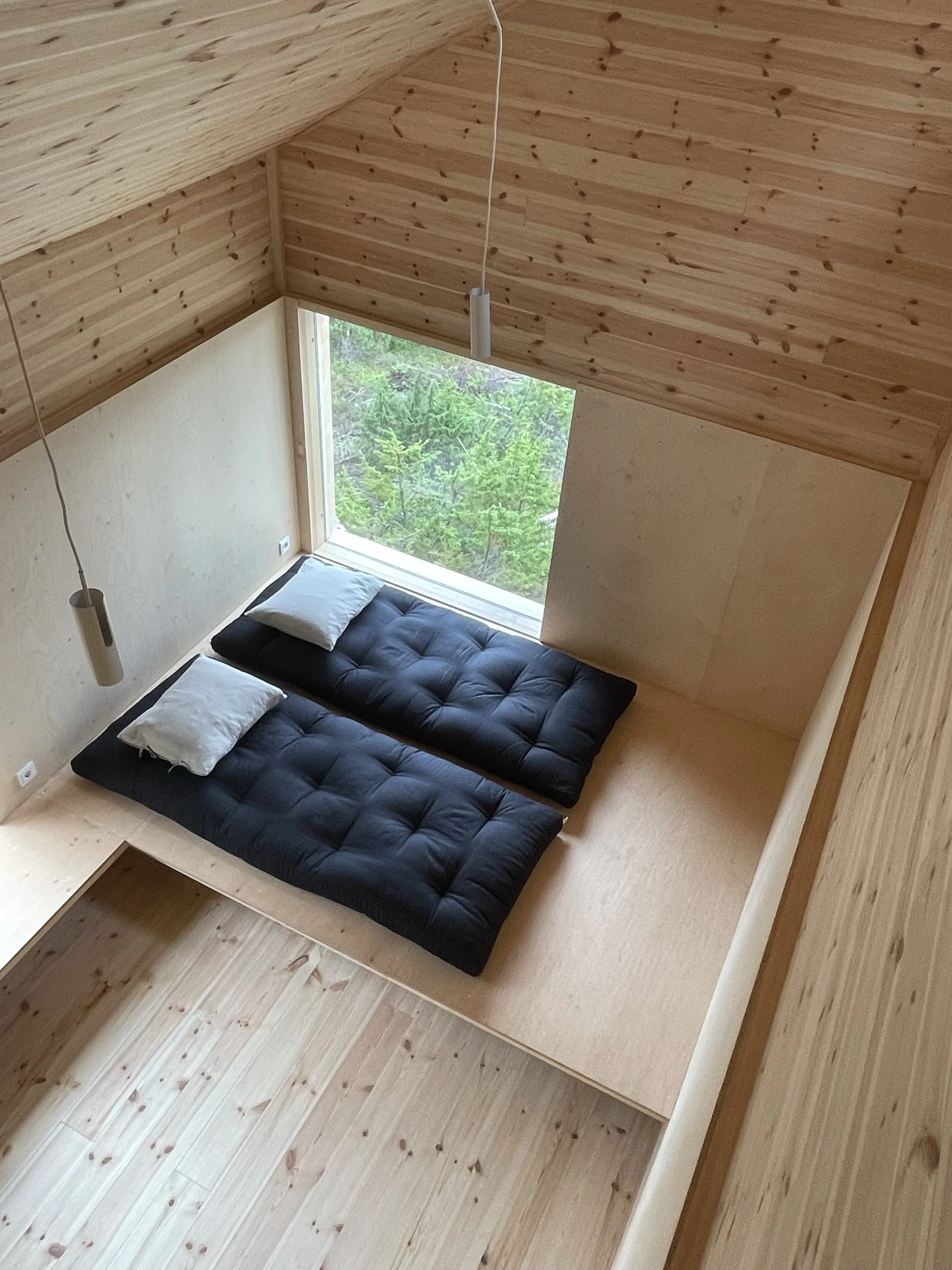 A minimalist bedroom with two black floor mattresses, each with a pillow, placed on a wooden platform with a large window overlooking green trees. The room has wooden walls and ceiling, with two hanging pendant lights.