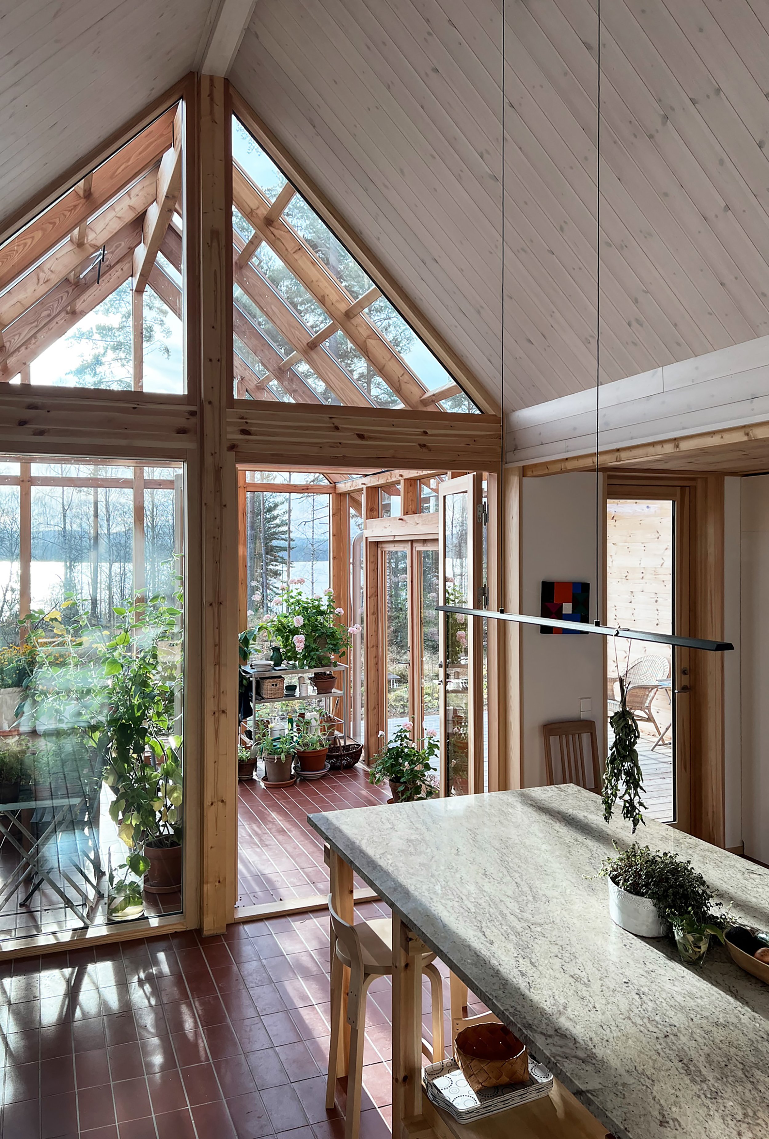 Interior of a sunlit wooden house with large glass windows and ceiling, displaying lush green outdoor landscape, potted plants on a balcony, and a marble kitchen island with hanging herbs and a small plant.