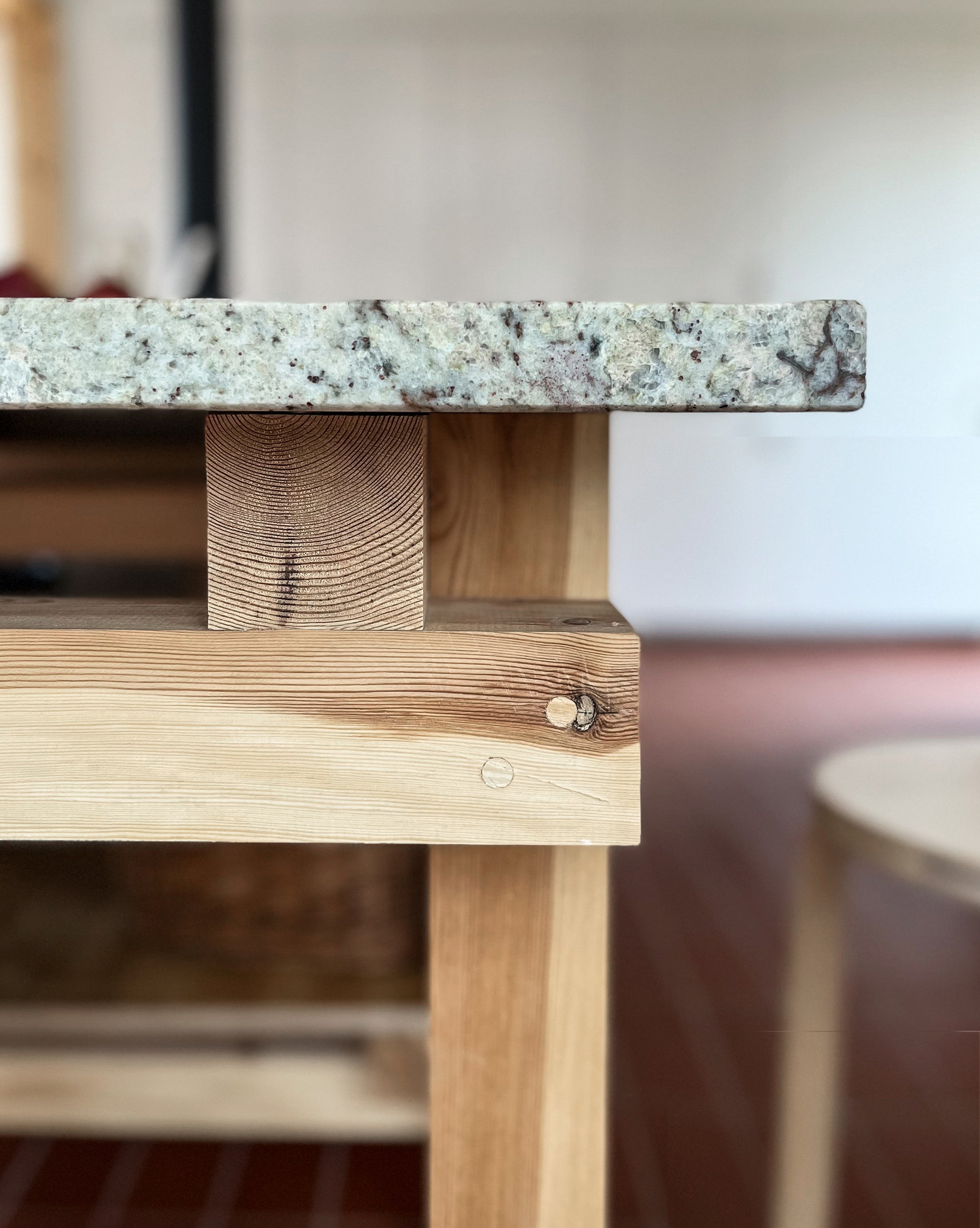 Close-up of a wooden table with a granite top, showing the edge of the table, details of the wood grain, screws, and supports, with a blurred background.