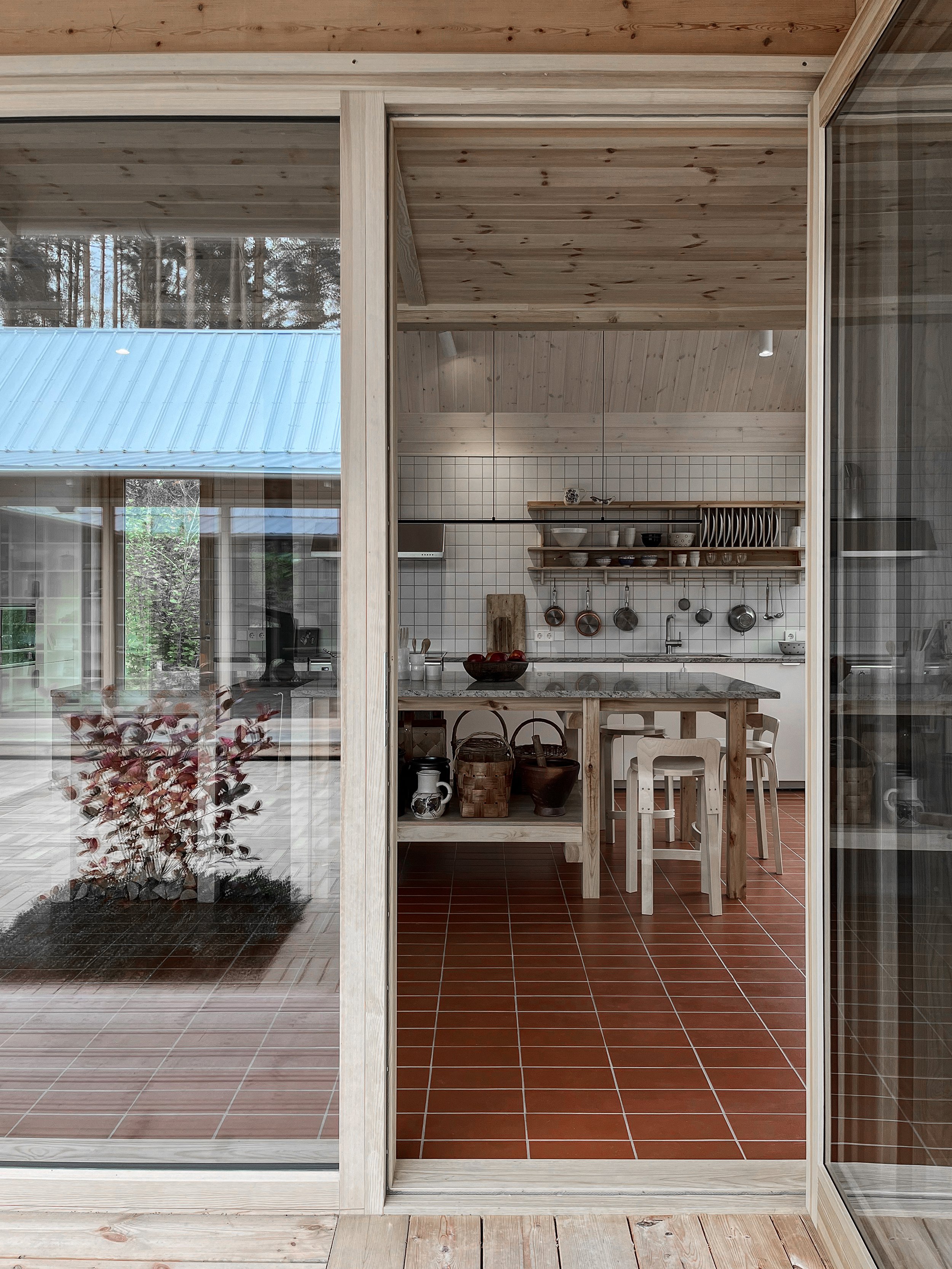 View of a cozy kitchen seen through a sliding glass door with a wooden frame. The kitchen features a dining table, chairs, open shelves with dishes, and hanging pots and pans. Tiled floor and wooden ceiling add warmth to the space.