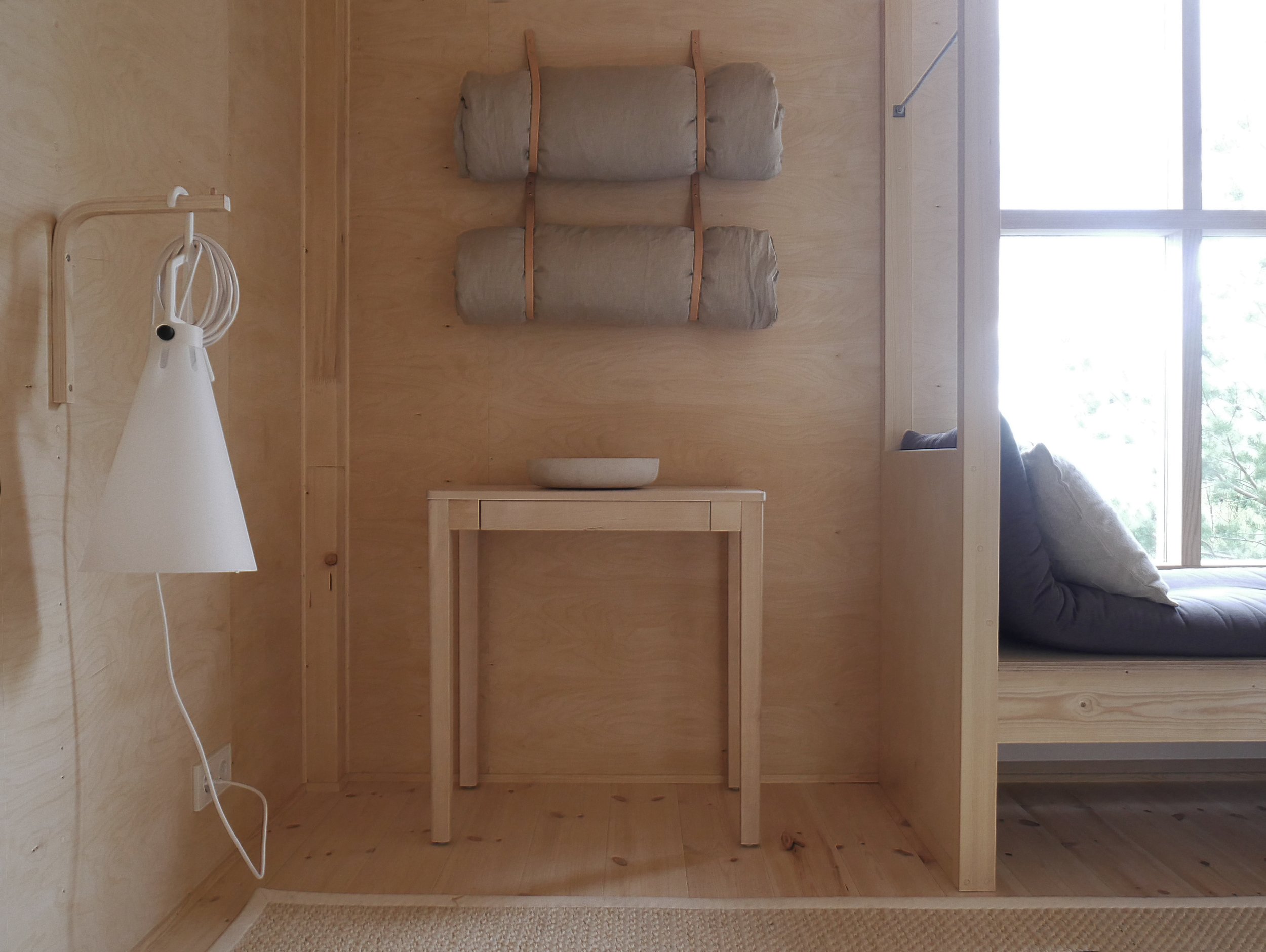 Minimalist interior with wooden walls and furniture, including a small table with a bowl, wall-mounted bedding, a hanging reading lamp, and a window seat with cushions.
