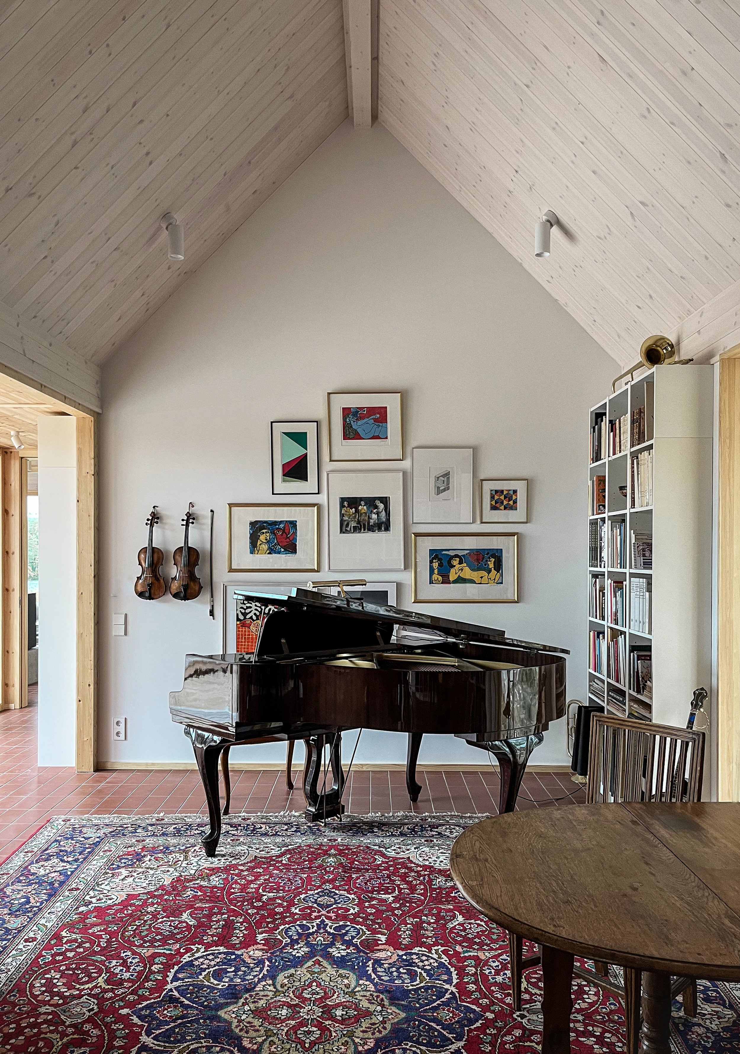 A cozy living room with a polished black grand piano, two violins hanging on the wall, framed artwork above the piano, a bookshelf filled with books, a wooden dining table, and a colorful patterned rug.
